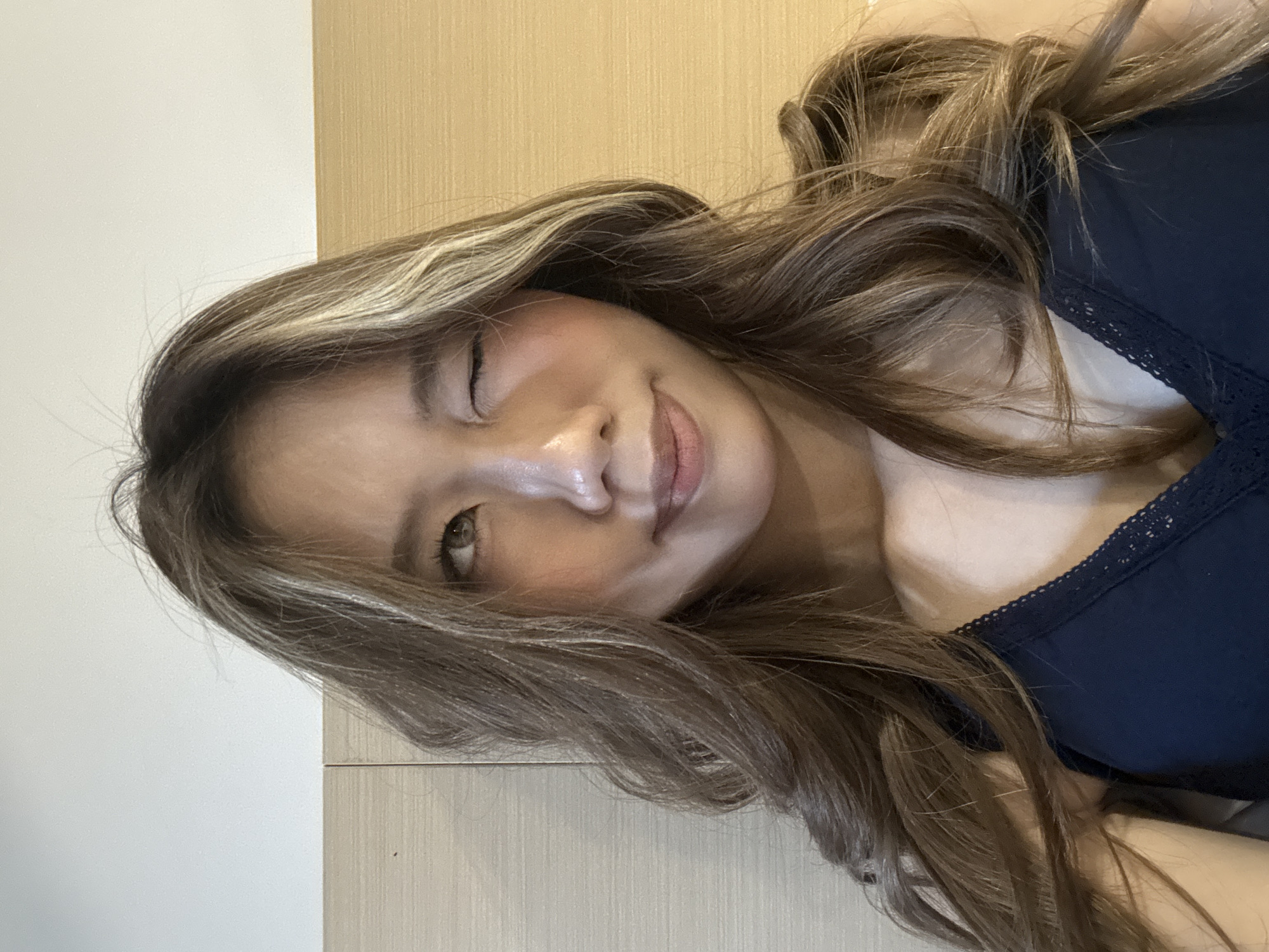 A young woman with long, wavy, light brown hair, featuring prominent blonde highlights, is captured in a close-up, rotated portrait. Her head is angled, suggesting she is lying down. She has an oval-shaped face with delicate features, including a small nose and full lips. Her left eye appears almost closed in a gentle wink or squint, while her right eye is partially open and looks towards the viewer. A soft, subtle smile plays on her lips, and a hint of blush on her cheeks adds to a playful and contented expression. She is wearing a dark navy blue top, possibly a tank top or camisole, adorned with delicate lace trim along the neckline. The background is split between a plain light cream-colored wall on the left and a vertical section of light wooden panelling on the right, indicating an indoor setting. The lighting is soft and even, highlighting her features and the texture of her hair without harsh shadows. The image portrays an intimate moment, likely a selfie, conveying a relaxed and amiable mood. There is no visible text. The location context for the image is Yuzhong District, China.
