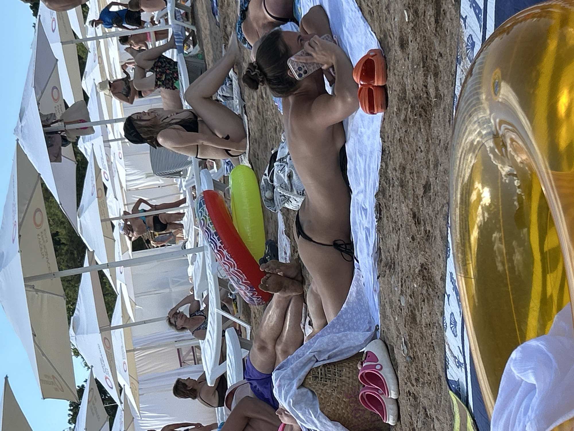 The image shows a beach scene in Gelendzhik, Russia, with several people relaxing on lounge chairs under white umbrellas. In the foreground, a woman is lying on a towel, using her phone, with orange slides nearby. To her left, a basket with a white cloth and pink slides is visible. In the middle ground, inflatable rings, a person in purple shorts, and their legs are present. Further back, other beachgoers are seen under umbrellas, with some in swimwear. The environment is a sandy beach with trees and the sky visible in the background.