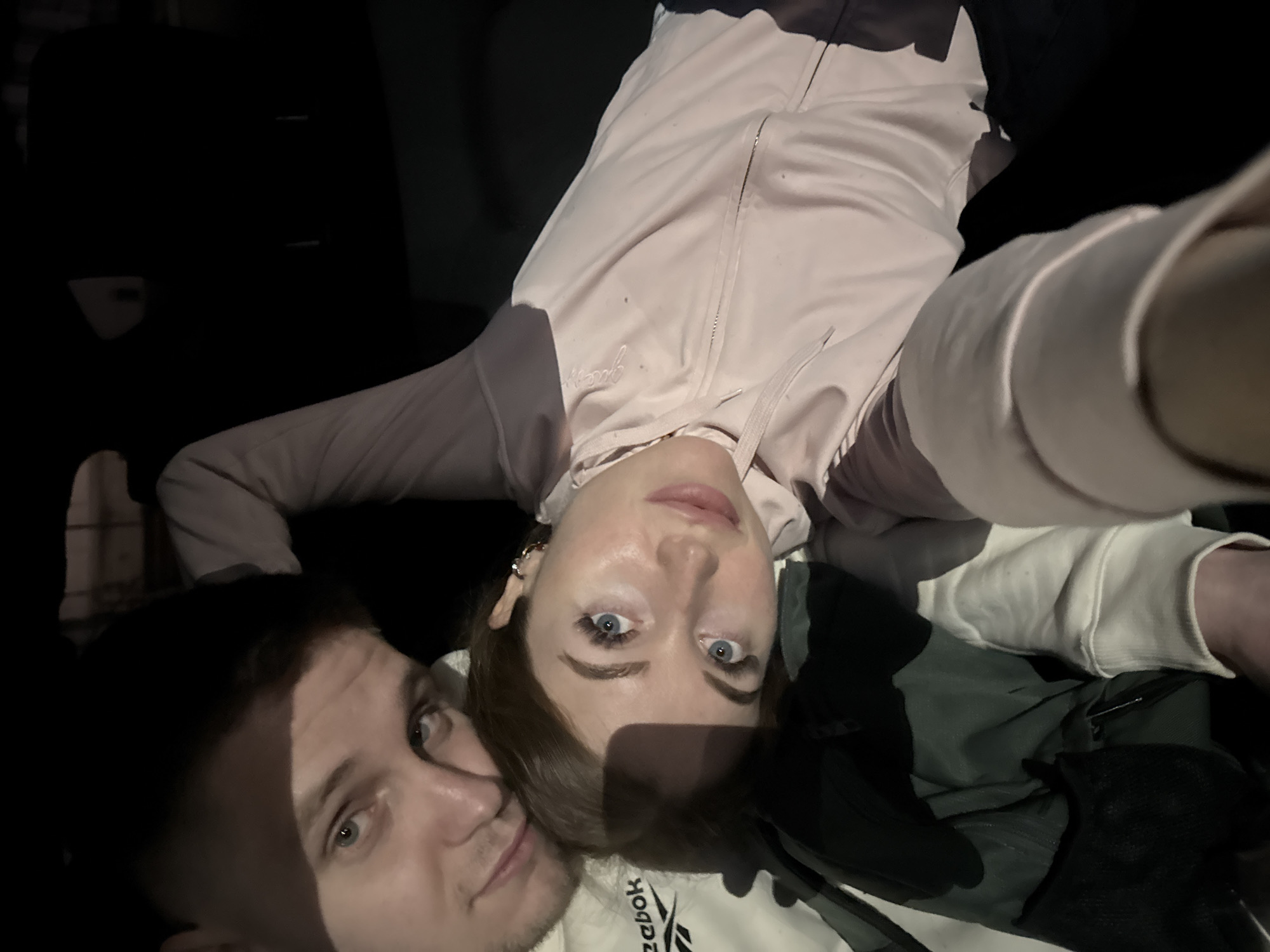 A low-angle selfie taken indoors shows a fair-skinned woman and a fair-skinned man. The woman is positioned in the upper right of the frame and wears a light pink jacket with gray sleeves. Her long brown hair is partially visible. The man is in the lower left and looks directly at the camera.
The background is predominantly dark. A black upholstered object is visible behind the woman’s jacket. A white Reebok sweatshirt is seen partially. The location is unknown.