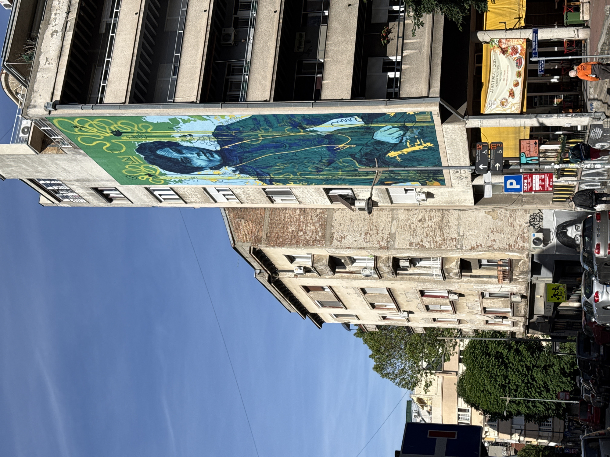 This image presents a low-angle view of a street scene in Belgrade, Serbia. The focus is on the architectural facade of a building featuring a large mural depicting a male figure in hues of blue and green, adorned with golden swirling lines. Adjacent to the building is another older structure with brickwork, adorned with air conditioning units.
The background showcases a light blue sky, with trees peeking in the lower right corner. Street signs and a banner for "AFIFA HALAL FOOD" are visible, along with parked cars and passersby on the sidewalk. The scene is bathed in daylight, suggesting it's daytime.