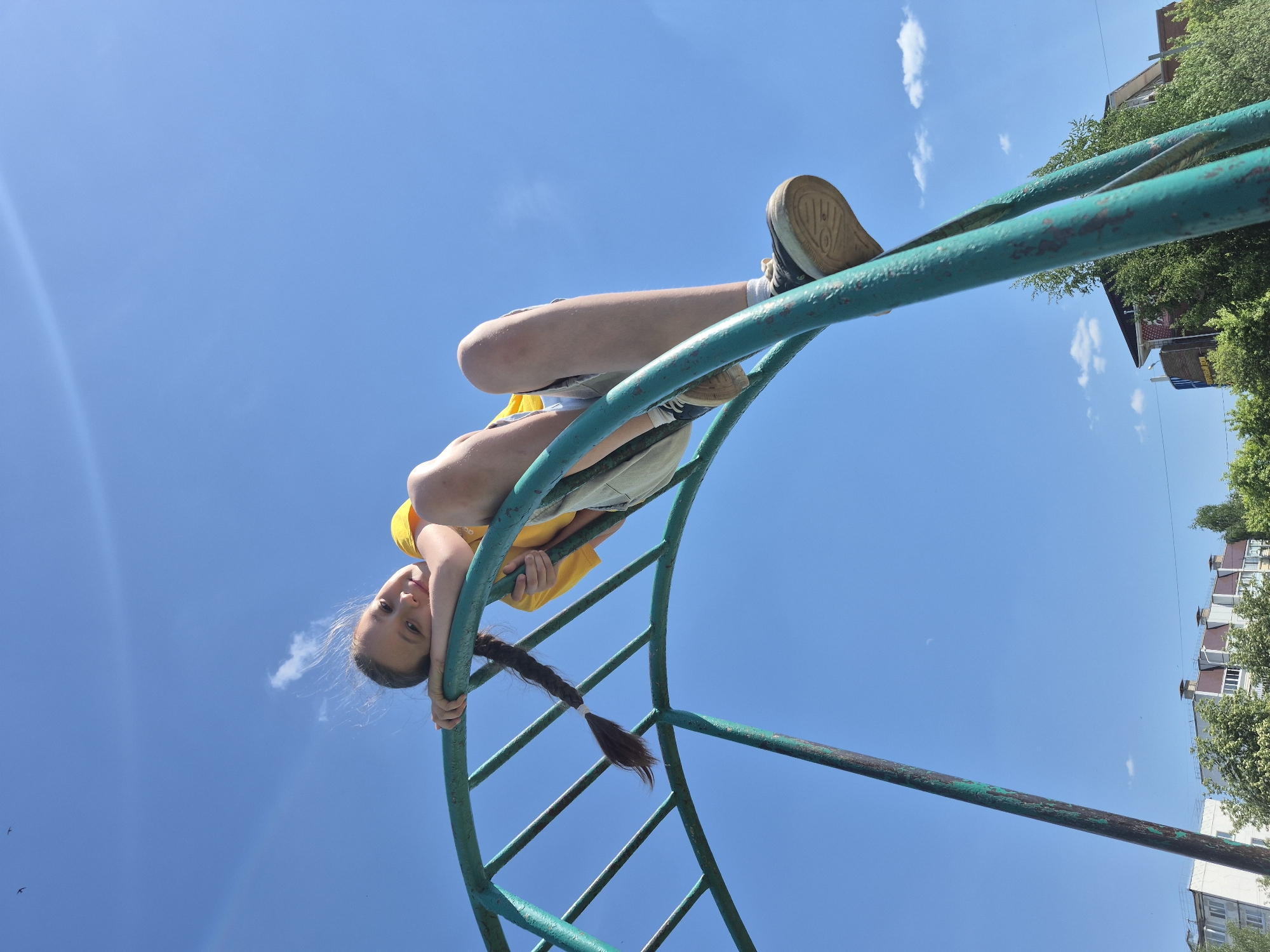 A young girl is hanging upside down from a playground structure in an outdoor environment. The structure, painted in green, consists of curved metal bars forming a climbing frame. The girl is wearing a yellow shirt and light-colored shorts. She is positioned at the lower end of the structure, with her shoes near the upper end. A building and trees are visible in the background, and the sky is blue with a few clouds.