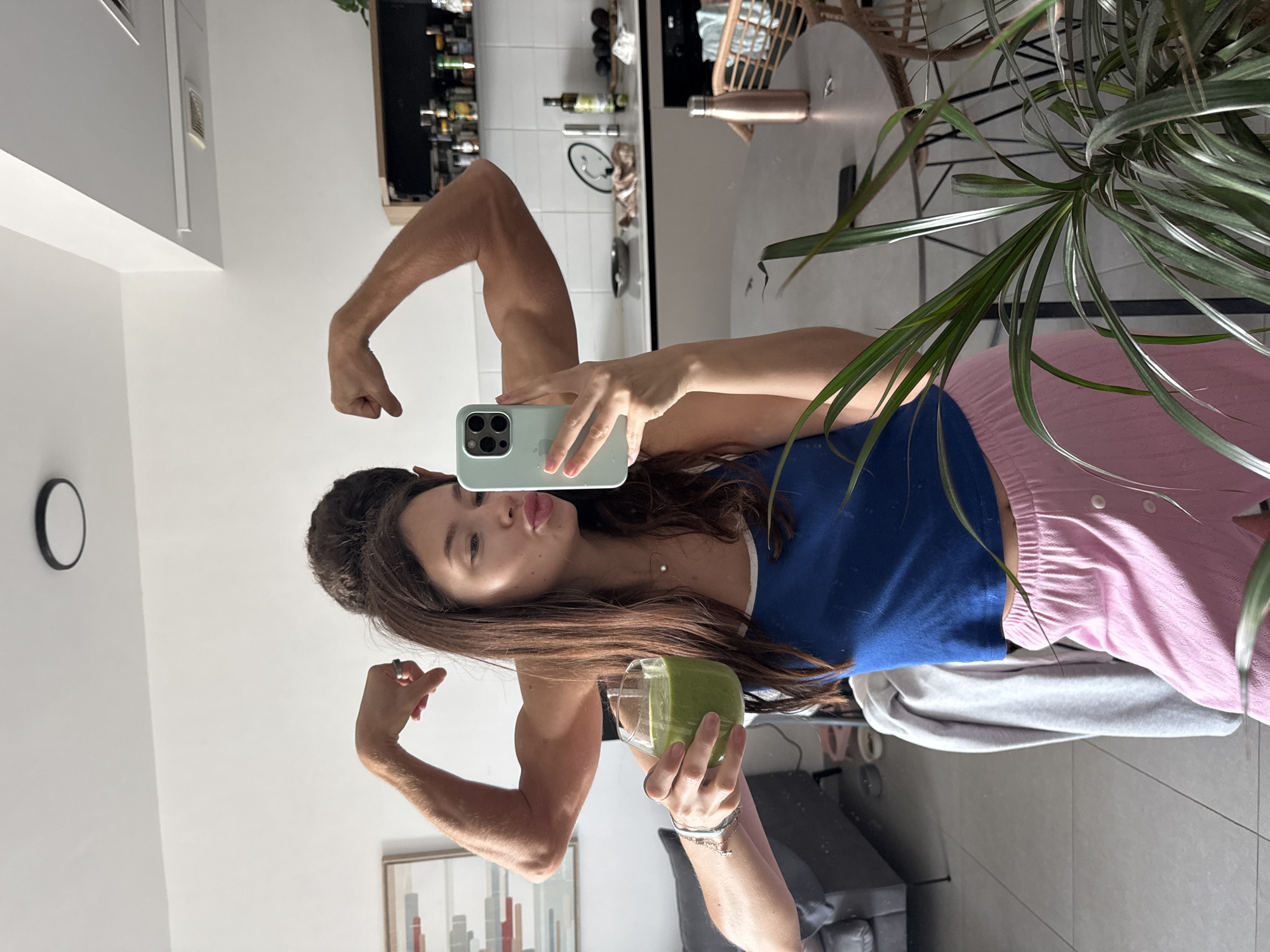 This mirror selfie captures a young woman posing playfully in an indoor residential setting, likely in Ramat Gan, Israel. She is seen flexing her biceps on both arms and making a "duck face" or kissy face towards the camera, conveying a confident and cheerful mood.
The woman has long, dark brown hair and is wearing a blue tank top with white trim and pink, textured pajama-style shorts or pants. On her left hand, she wears a silver ring, and on her right wrist, she has multiple silver bracelets. She holds a light blue iPhone, which appears to be an iPhone 12, 13, or 14 Pro/Pro Max due to its triple camera system, in her left hand, using it to take the photo. The word "iPhone" is visible on the back of the phone case. In her right hand, she holds a clear glass filled with a vibrant green smoothie, suggesting a focus on health and wellness.
The reflection in the mirror reveals a modern and well-appointed interior. Behind the woman, a kitchen area is visible with shelves stocked with various bottles, likely cooking oils and spices. Further right, a dining area is reflected, featuring a round dining table, a light-colored woven rattan chair, and a copper-colored metallic water bottle resting on the table. A large potted plant with long, thin green leaves partially frames the right side of the image, adding natural decor. In the background, a minimalist black-framed clock hangs on a white wall, and a framed abstract artwork with vertical colorful blocks is partially visible. The general lighting suggests it is daytime.