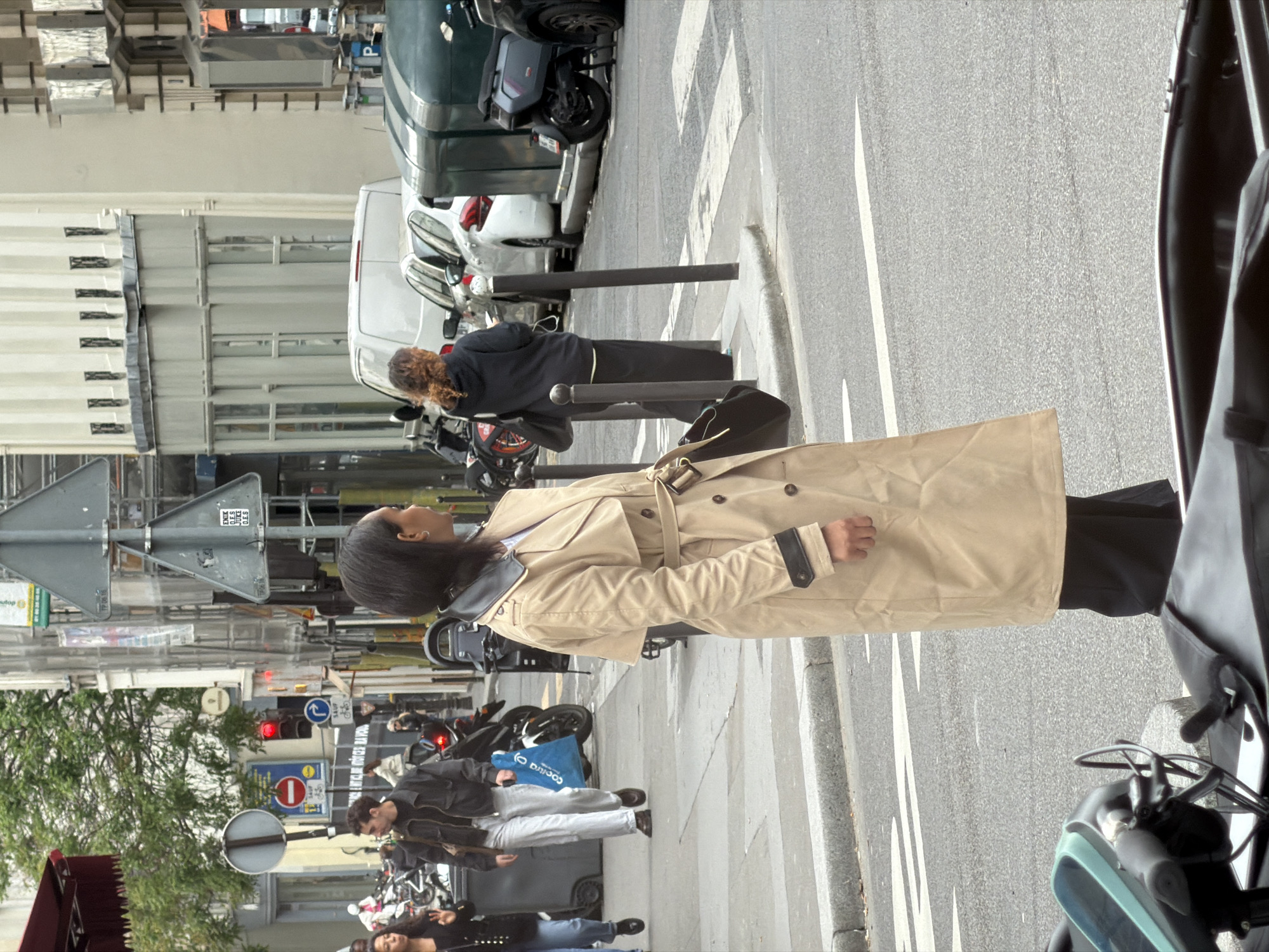 The image depicts a street scene in Paris, France. A woman in a tan trench coat and black pants stands on a zebra crossing, looking towards a street sign. There are people walking and standing on the sidewalk, and parked motorcycles line the street. Buildings with light-colored facades are visible in the background. Cars and vans are parked alongside the street. A woman with curly hair wears a black jacket and dark pants, standing near a black bag.