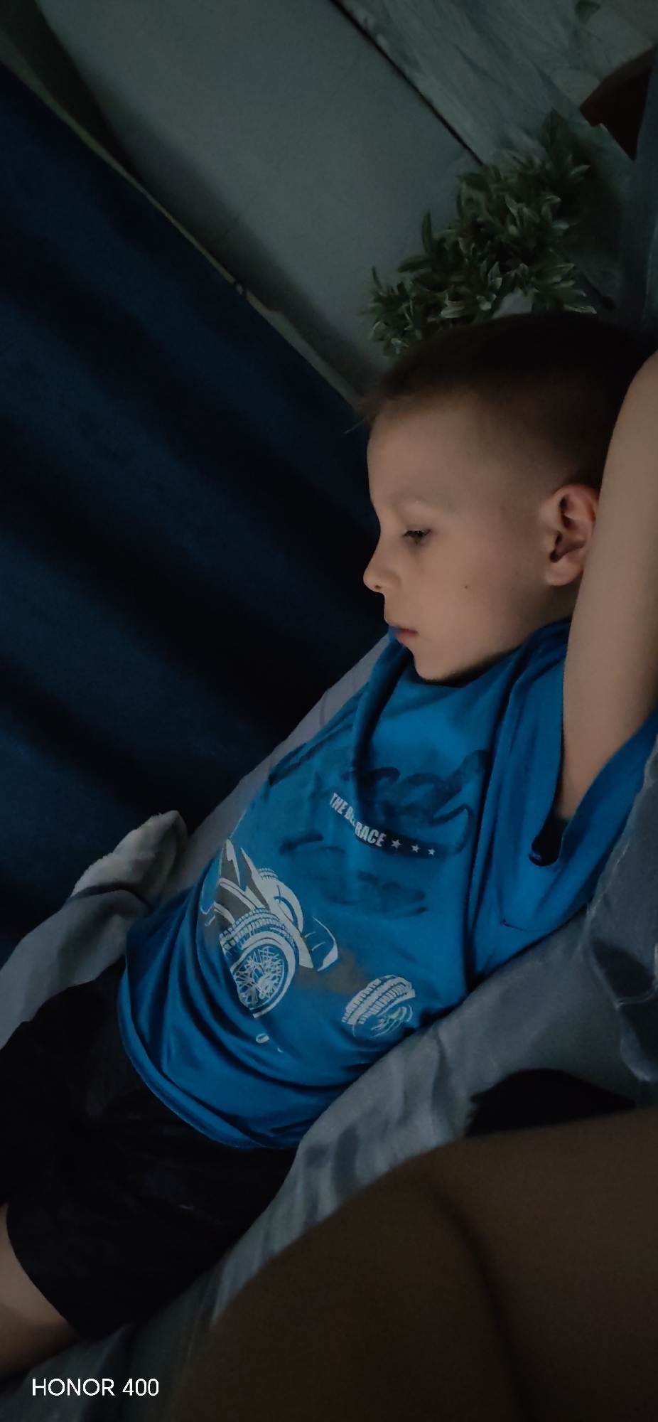 A young child is lying on a bed. He has short, light brown hair and is wearing a bright blue t-shirt with a graphic design featuring a race car. The child's arm is visible next to his head. The background includes a dark blue curtain and a plant with green leaves.
There is no visible information in this image to ascertain it being in the city of Noviy, Russia.