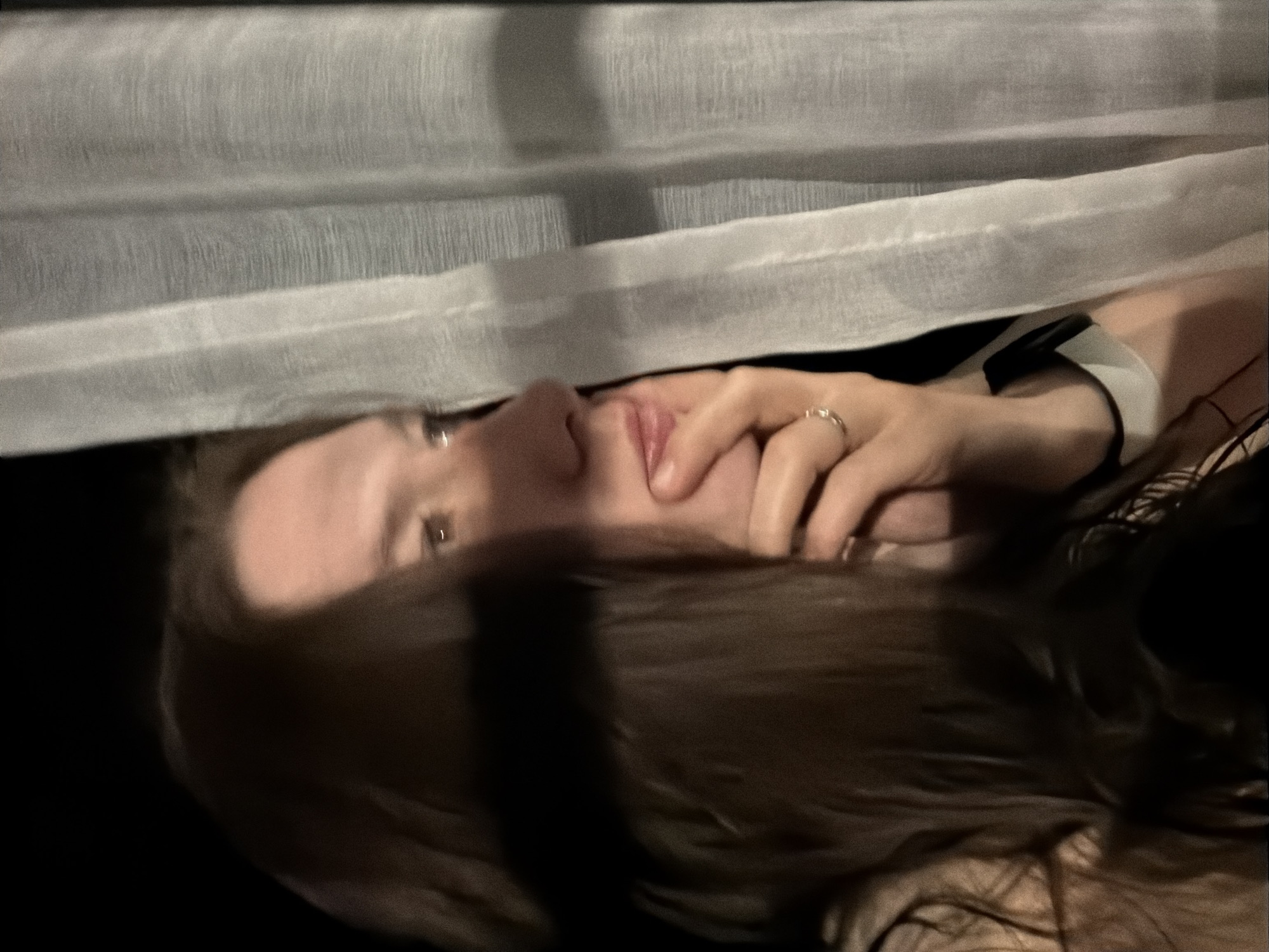A medium shot shows a person looking upward from underneath a white translucent fabric, possibly a curtain or sheet. The person has fair skin, light eyes, and brown hair. Their hand, adorned with a ring, touches their face near the mouth. The background appears to be dark. The location, Staraya, Russia, cannot be verified from the visual information provided.