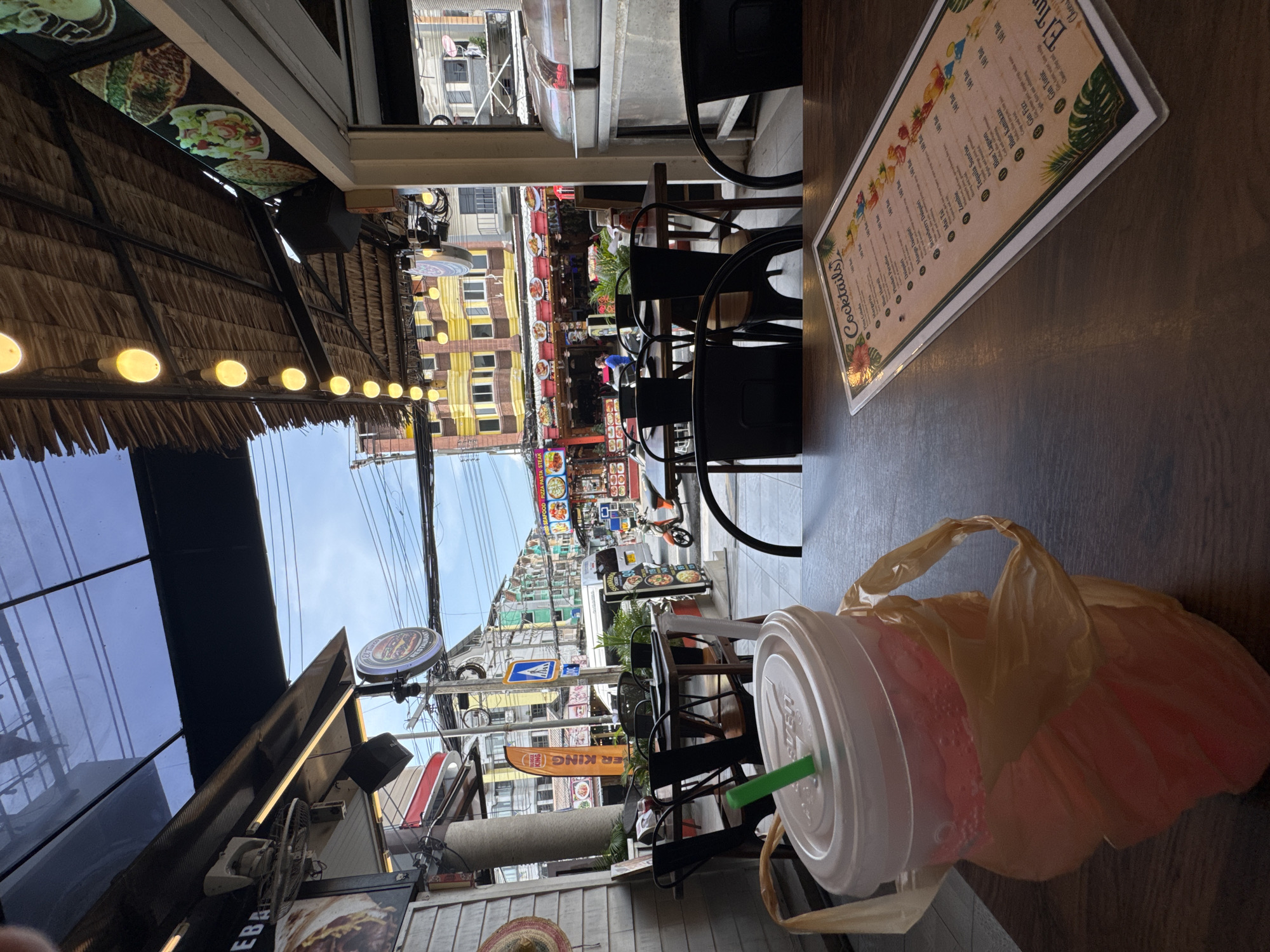 A frontal, eye-level shot captures a street scene in Karon, Thailand, with a focus on a table in the foreground set for a meal. On the table rests a pink beverage in a clear plastic cup with a white lid and green straw, partially obscured by a pale orange plastic bag. To the right, a cocktail menu is laid open, displaying drink names and prices. Beyond the table, outdoor seating with dark chairs and tables is visible, leading to a bustling street. Shops line the street, adorned with colorful signage, including "Burger King." The sky above is a clear blue with a few clouds, and the scene is illuminated by natural light and overhead string lights. A motorcycle is parked on the street, and a few people can be seen walking in the background. The architecture suggests a tropical setting, with thatched roofs and open-air structures.
