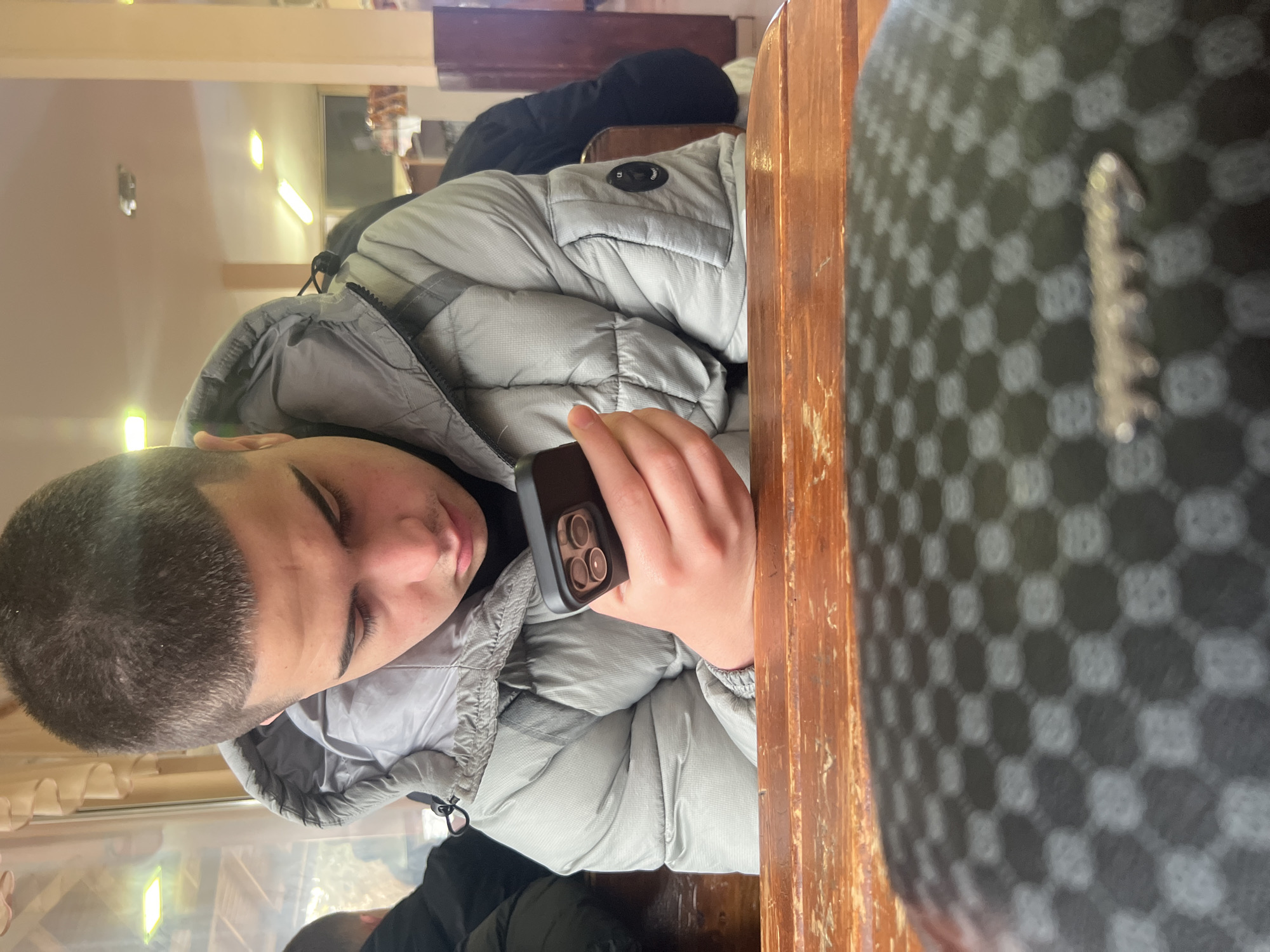 A young male is seated at a wooden table, holding a black cell phone in his right hand. He has short dark hair and is wearing a light grey puffer jacket. In the foreground, a checkered object obscures part of the scene. The setting appears to be indoors, with light-colored walls and light fixtures visible in the background. The location is Asenovgrad, Bulgaria, though the specific details of the location (e.g., name of establishment) are not discernible from the image.