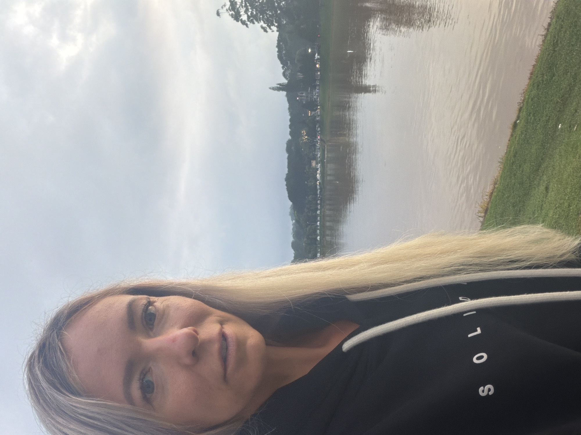 A blonde woman wearing a black hoodie with white drawstring and white lettering on the chest stands on a grassy bank facing a large body of murky water. The woman's face is partially visible, with her long blonde hair falling over her right shoulder. Behind her, across the water, is a treeline with some buildings and lights visible amongst the foliage. The sky is overcast. This appears to be Xuan Huong Lake in Da Lat, Vietnam.