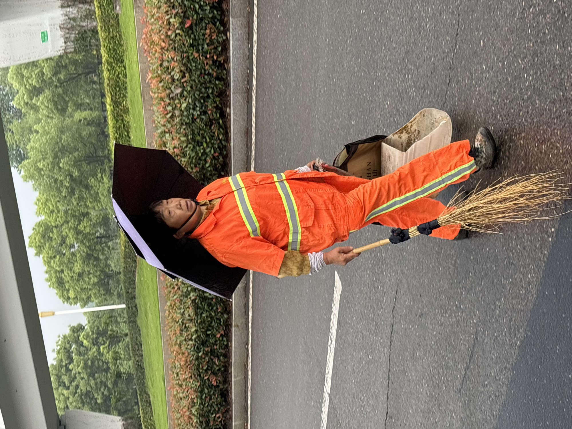 The image shows a woman in an orange uniform standing on a gray asphalt road with a broom in her right hand and a black umbrella held above her head with her left hand. The road is next to a green hedge. She has a cardboard container behind her right leg. The words "HAN SHAN" are visible on the side of the cardboard container. The scene is likely in the city of Jiangjin, China based on the text on the cardboard container.