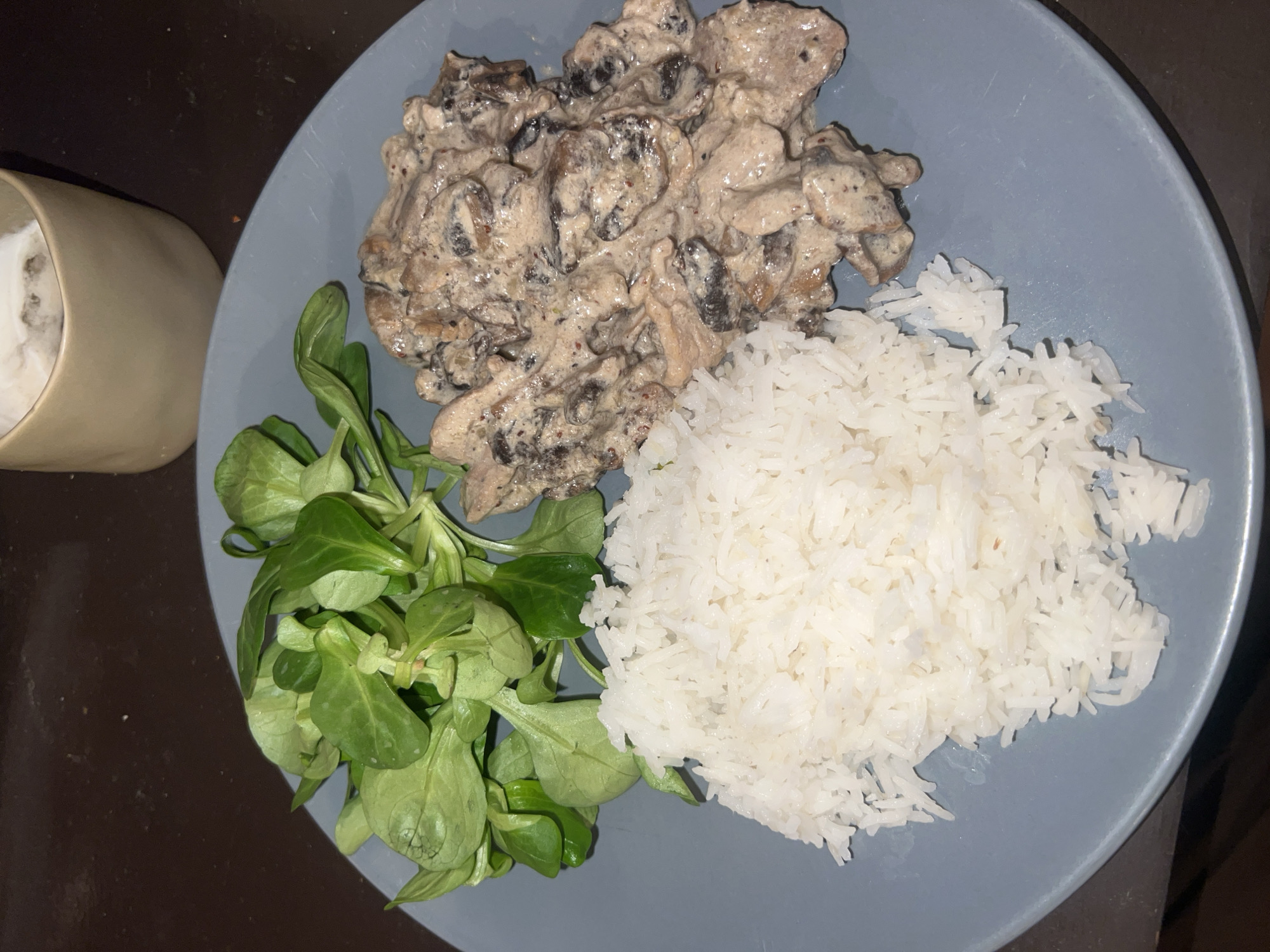 A plate of food is presented. The plate is light blue and circular. The food consists of a pile of white rice, a clump of green salad, and a mound of creamed mushrooms with pieces of meat. A beige mug sits to the left of the plate. The background is a dark brown surface. The location is not discernible as Paris, France.