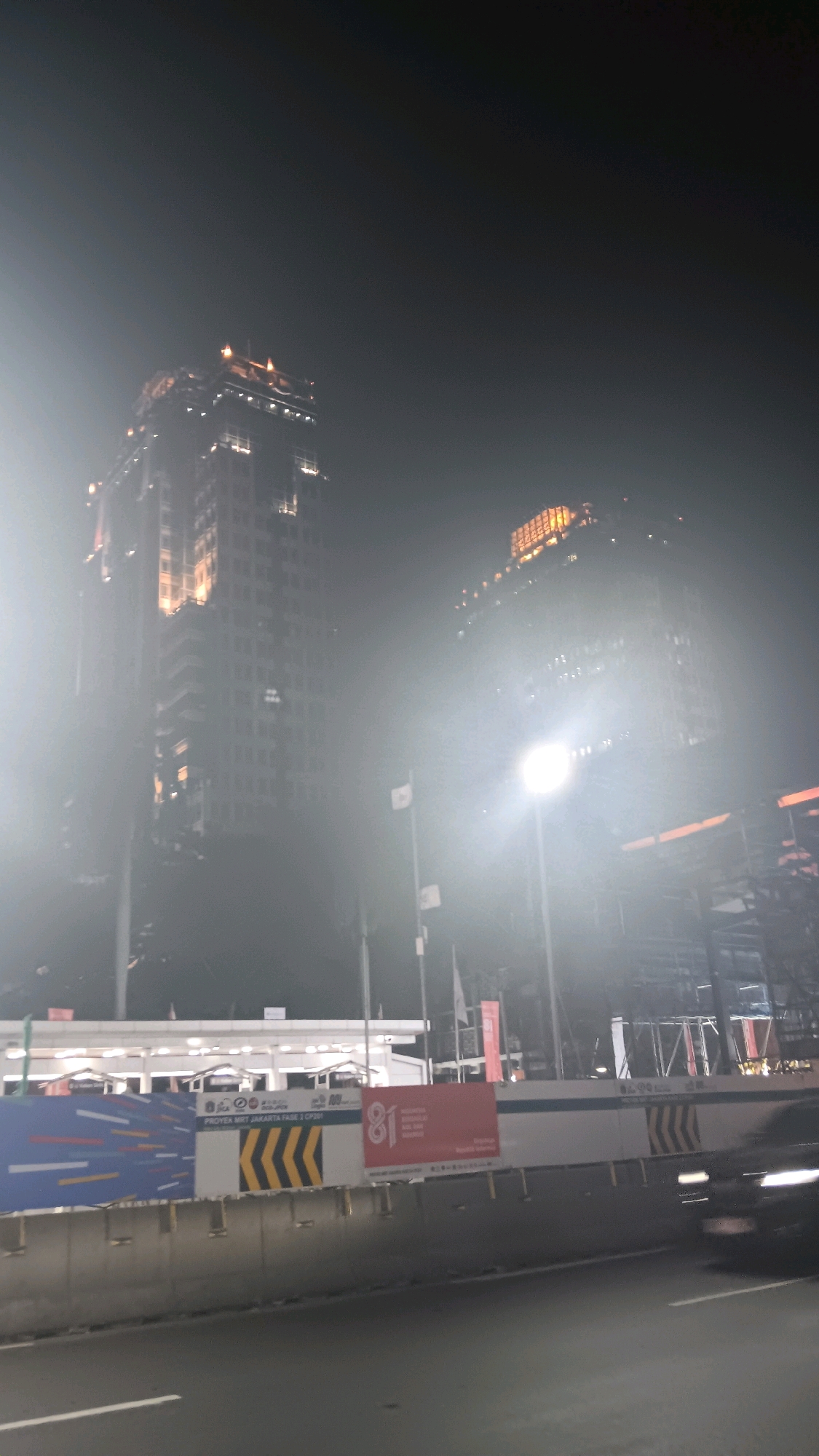 The image shows a nighttime view of a construction site in Central Jakarta, Indonesia. In the background, two tall buildings are visible, illuminated by lights. One building on the left appears to be a completed structure with lights on its upper floors and along its facade. The building on the right is also lit up, with some parts appearing to be under construction.
In the foreground, there is a road with a concrete barrier. Behind the barrier, there is a long white structure with several illuminated openings, possibly a temporary shelter or part of the construction. Banners with graphics and text are displayed on the barrier. One banner features a large number "81" in red, and another banner displays colorful geometric patterns and the text "PROYEK MRT JAKARTA FASE 2 CP201". There are also several flags on poles.
A vehicle is visible on the road, partially obscured by motion blur, suggesting it is moving. The overall atmosphere is hazy, possibly due to fog, dust, or light pollution, which diffuses the light sources and softens the details of the scene. The sky is dark, indicating it is nighttime.