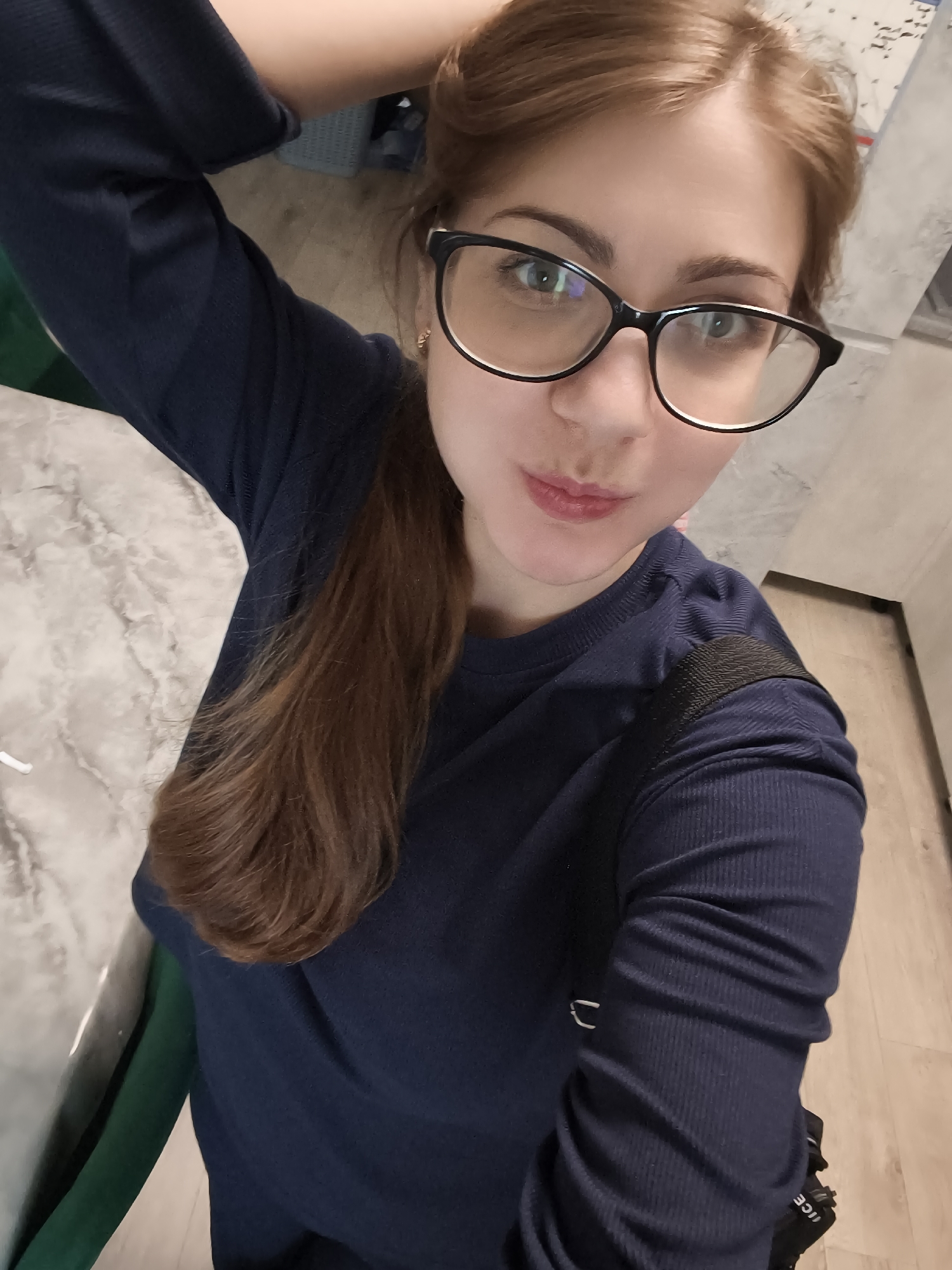 A young woman with long, light brown hair pulled into a ponytail is taking a selfie. She is wearing a dark blue, ribbed long-sleeved shirt and black-rimmed glasses. Her right arm is raised behind her head, and she is looking directly at the camera with a slight pout. The background shows a marble-like countertop, a green object partially visible, and a wooden floor. The scene appears to be indoors, likely in a home setting. The lighting suggests it could be daytime or artificial lighting indoors. The woman's expression is playful and relaxed. No visible text is present in the image. The location context is Novaya Usman', Russia, but there are no specific visual cues within the image to confirm this location.