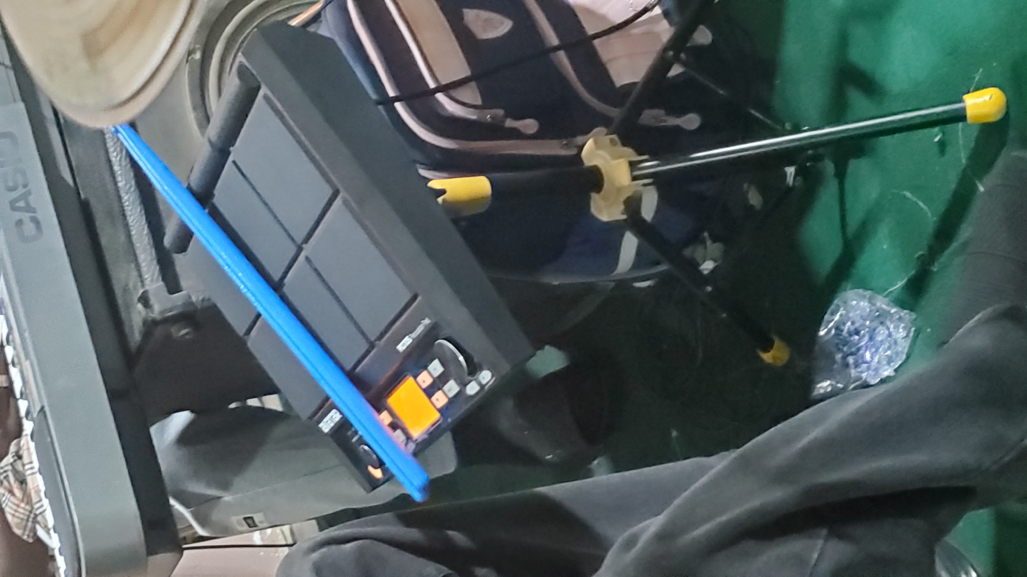 The image depicts a close-up, eye-level view of a music production setup, likely in a residential or studio environment in Jalingo, Nigeria. The scene is dominated by a black drum pad controller, which is tilted upwards and angled towards the viewer. A bright blue drumstick rests horizontally across the top of the drum pad, obscuring some of its surface. The drum pad features several illuminated orange buttons and a small screen displaying orange text.
Behind the drum pad, a portion of a grey Casio keyboard is visible, suggesting a broader musical setup. To the right, a dark blue and white backpack rests on the floor, partially obscuring the legs of a black tripod stand, which is supporting some unseen equipment. The tripod has yellow accents on its feet and at the top. The floor appears to be covered in a dark green carpet or matting, and in the lower left quadrant, a person's legs, clad in dark grey trousers, are visible, implying their presence in the scene. A crumpled plastic bag containing small blue objects is also on the floor near the person's legs. The overall lighting is dim, with the primary illumination coming from the illuminated elements of the drum pad.