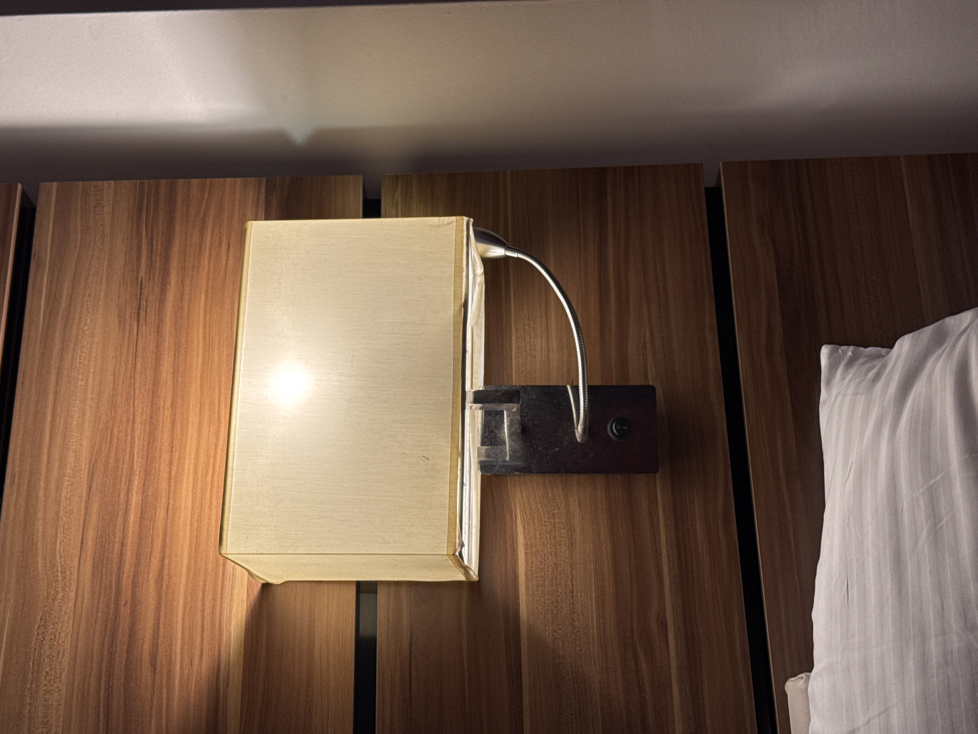 The image shows a bedside lamp mounted on a wooden headboard. The lamp has a rectangular, off-white fabric shade with a metallic base and a flexible gooseneck arm. The headboard is made of horizontal wooden panels with a warm, reddish-brown hue. Below the headboard, a white pillow with subtle grey stripes is partially visible. The scene appears to be indoors, likely in a bedroom. No people or specific actions are depicted. The location of Aksu, Turkey is not visually determinable from the image.
