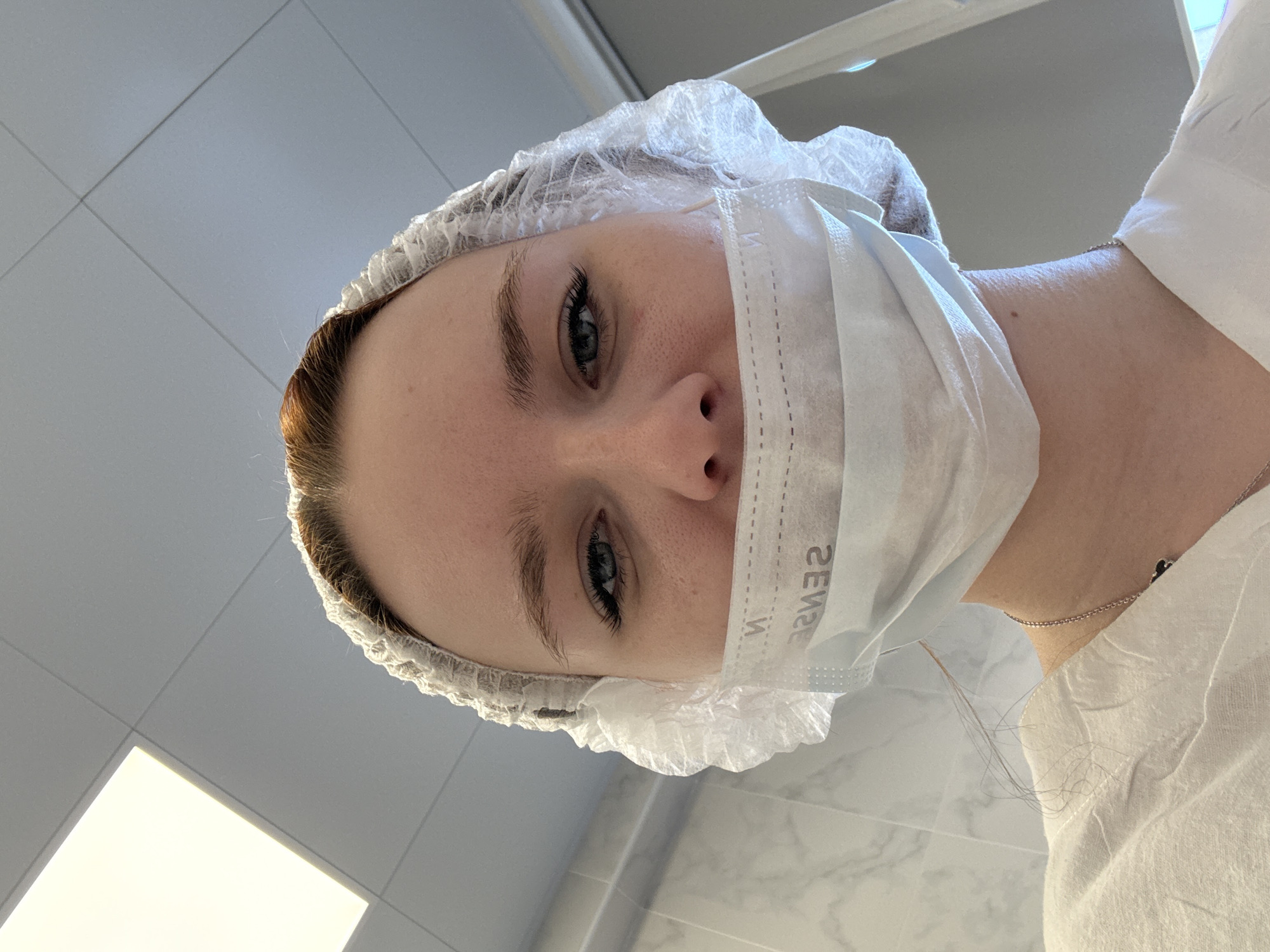 A person with light skin, blue eyes, and reddish-brown hair is wearing a disposable white hair net and a white surgical mask that covers their nose and mouth. The mask has the word "SENSE" printed on it in reverse. The person is wearing a white top. The background consists of white tiled walls and a fluorescent light panel. The location is not discernible in the image. The city of Saint Petersburg, Russia, cannot be confirmed from the details in the picture.