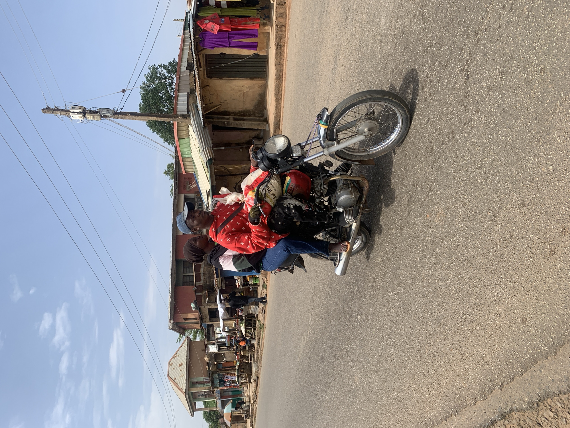 Two people ride a motorcycle on a paved road in Gura Top, Nigeria. The motorcycle is old and appears to be well-used. The driver wears a red shirt and a blue cap, while the passenger wears a head covering. The background consists of buildings with corrugated metal roofs and a wooden utility pole with wires. Clothing hangs from a line in front of one of the buildings. The sky is pale blue with a few wispy clouds. The road is asphalt.