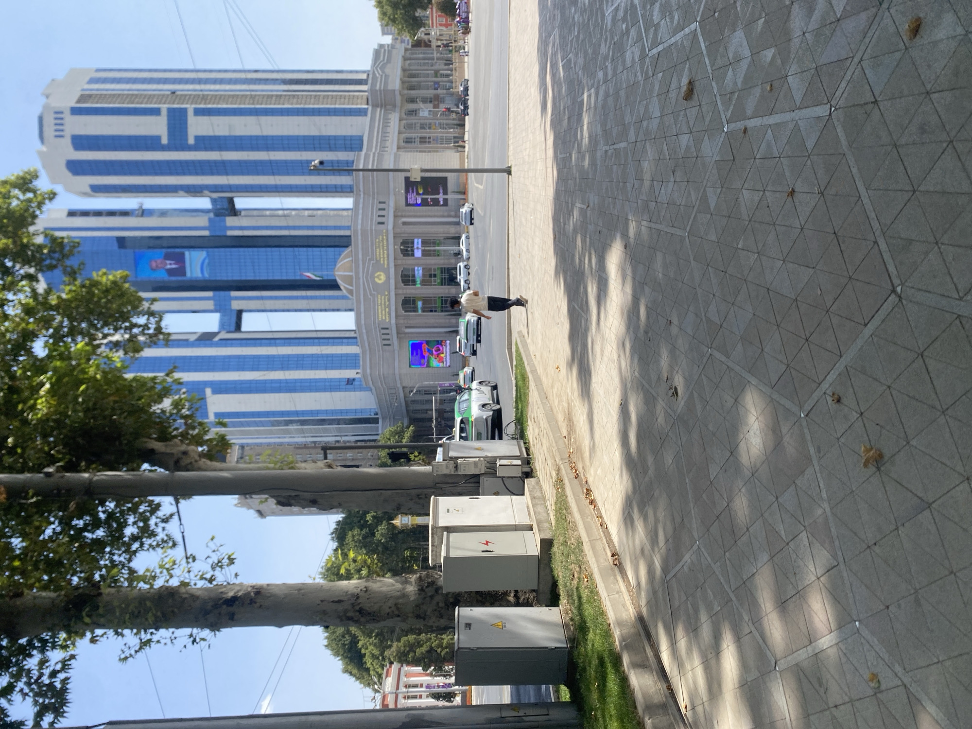 A street scene in Dushanbe, Tajikistan, features a modern, tall building with blue and white vertical accents. The building has a large banner displaying a portrait of a man in a suit. In front of the building is a wide, paved sidewalk and road. A person is seen walking away from the camera on the sidewalk, dressed in a light-colored shirt and dark pants. Several utility boxes are lined up on the side of the sidewalk, some with warning symbols. Cars are visible on the road, including a white car with a green stripe. Trees with green foliage are present along the street. The sky is clear and blue.