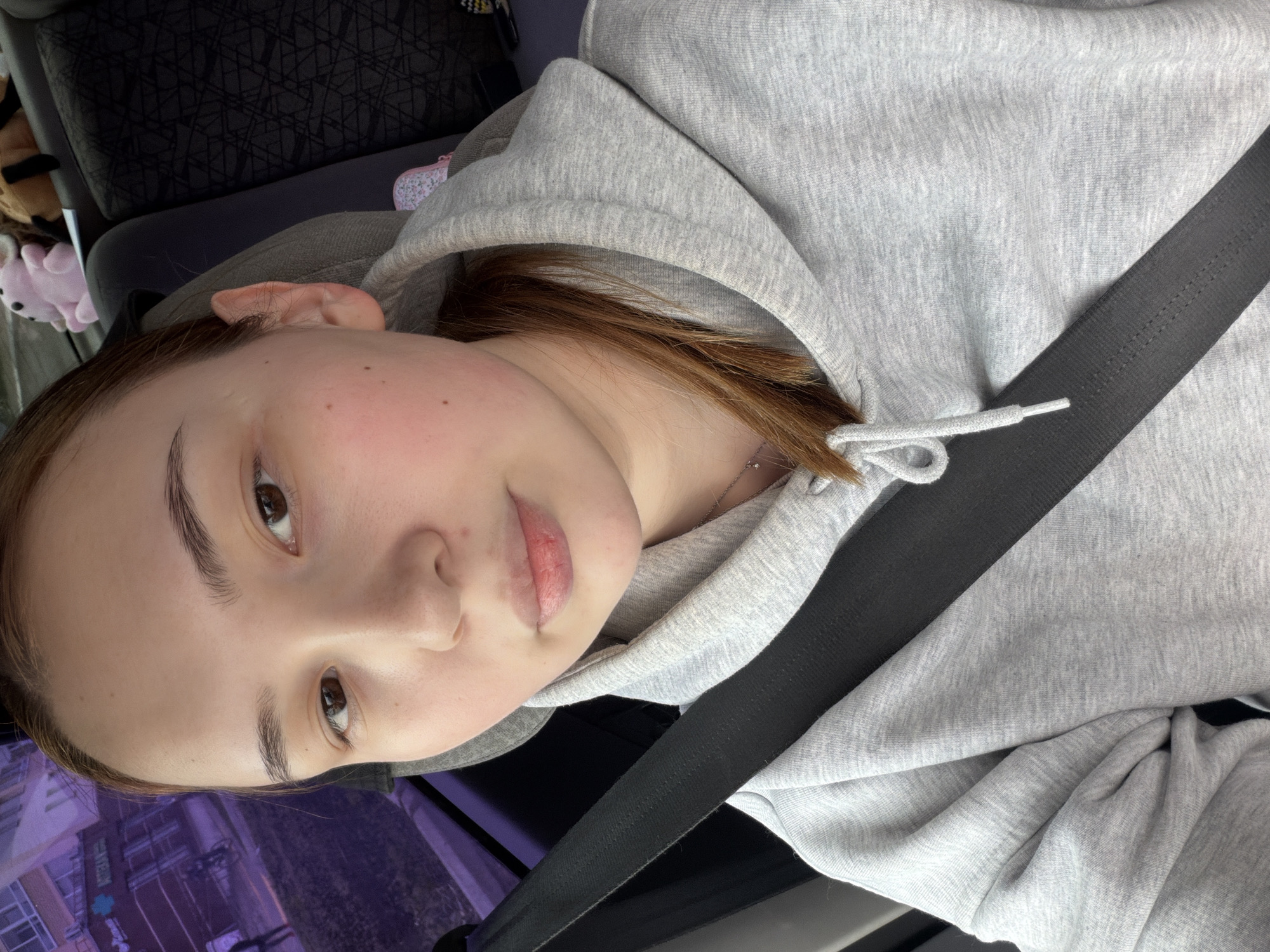 A young woman is photographed indoors, likely in a vehicle, based on the presence of a seatbelt across her torso. She is wearing a grey hooded sweatshirt. Her hair is light brown. Her skin tone appears fair. The background is blurred, partially revealing what appears to be a building with a purple hue reflected in the glass. A partial view of items possibly including toys and bags is seen in the upper left. While the location cannot be definitively determined to be Chaikovsky, Russia, based solely on the provided image, the background suggests an urban environment with buildings. The subject is looking directly at the camera.