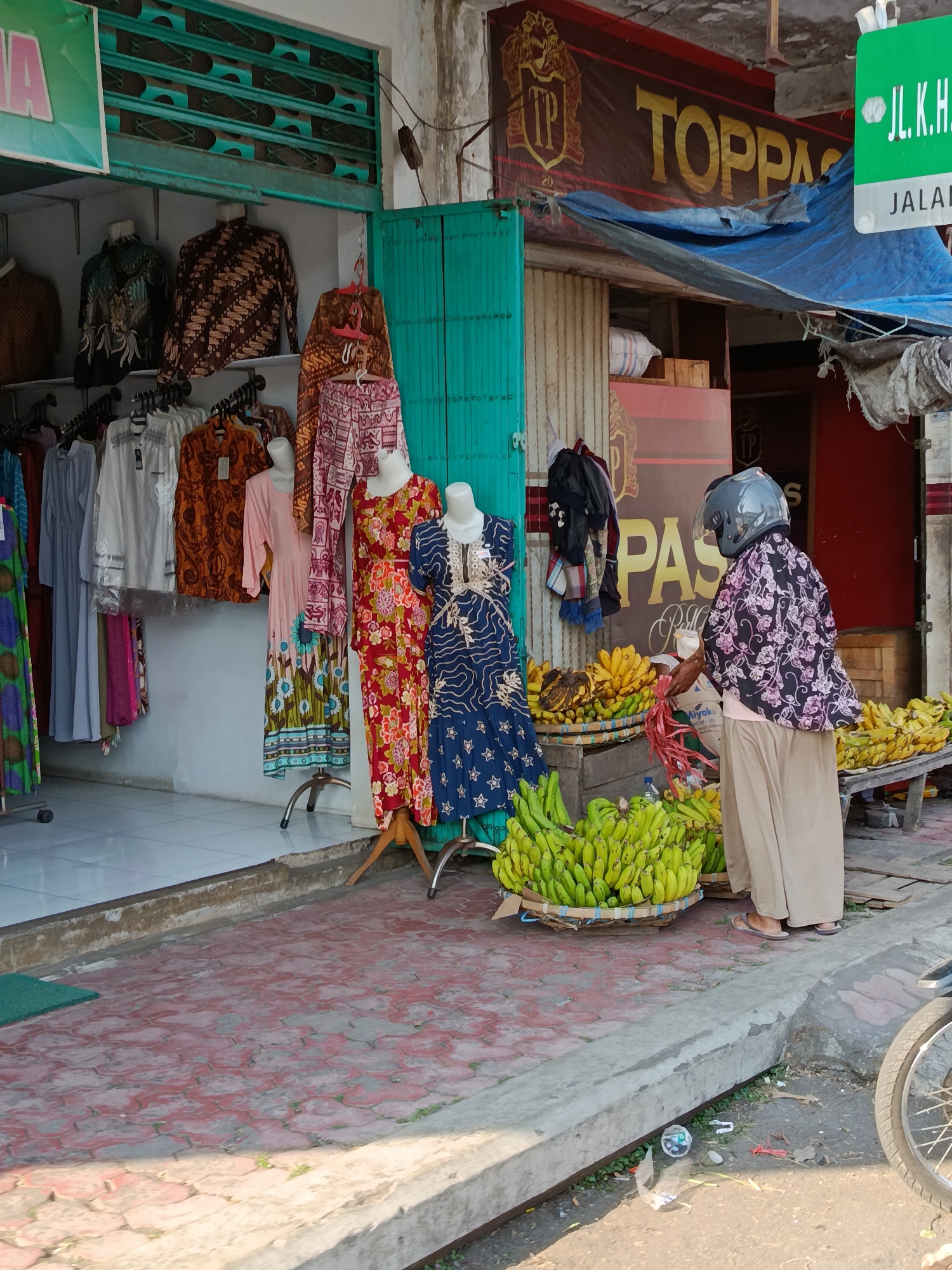 The image depicts a street scene in front of a shop in Bondowoso, Indonesia. A woman wearing a patterned shirt, light-colored pants, and a helmet is standing by a display of bananas. She appears to be interacting with the fruit. The shop has various clothing items, including batik shirts and dresses, displayed on mannequins and racks. Baskets overflowing with bananas are arranged on the sidewalk in front of the shop. The ground is paved with interlocking red bricks. In the background, a banner with the text "TOPPAG" is visible. The environment suggests a bustling market or commercial area.
