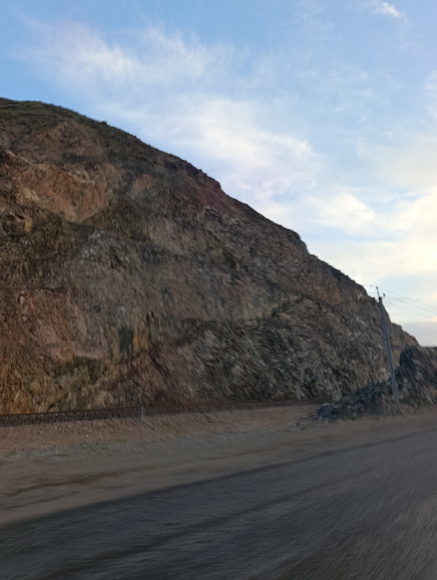The image depicts a mountainous landscape with a road in the foreground. A steep, rocky hillside dominates the background, with visible strata and varying shades of brown and gray. A power line pole and wires are situated on the right side of the frame, near the crest of the hill. The sky above is a pale blue with scattered wispy clouds. The foreground shows a paved road with some blurry motion evident, suggesting it was taken from a moving vehicle. A narrow band of dry grass and gravel separates the road from the base of the hill, where what appears to be a railroad track is visible. The scene is set in the city of Semiz Bel, Kyrgyzstan.