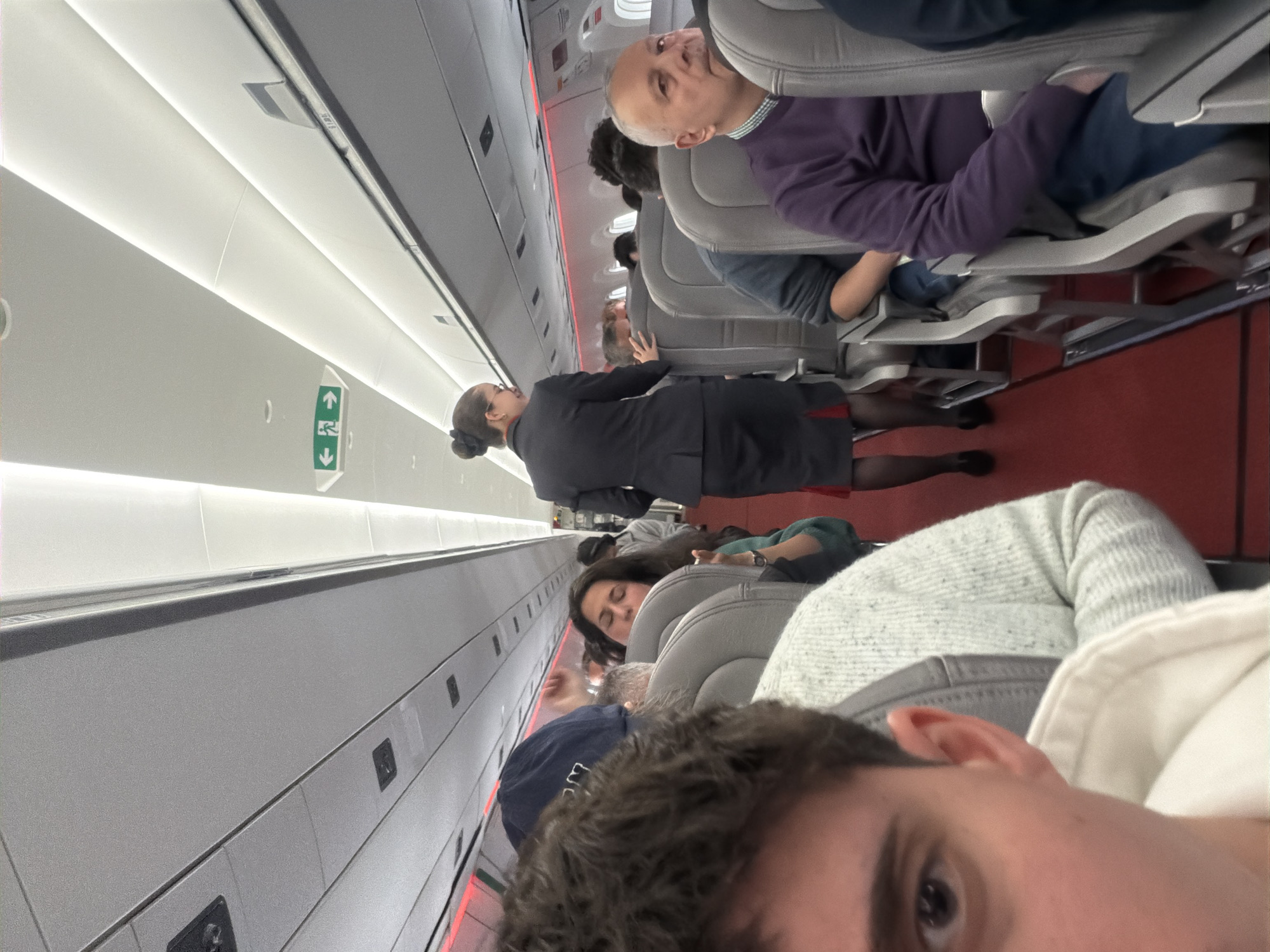 The image shows the interior of an airplane. An aisle divides rows of seats filled with passengers. A flight attendant in a dark uniform walks along the aisle. The seats are grey and the floor is red. The interior walls are light grey and overhead compartments are visible. The presence of a green emergency exit sign suggests the location is on an airplane, not in Sofia, Bulgaria. The camera is positioned in one of the seats, and the person holding the camera is visible in the foreground.