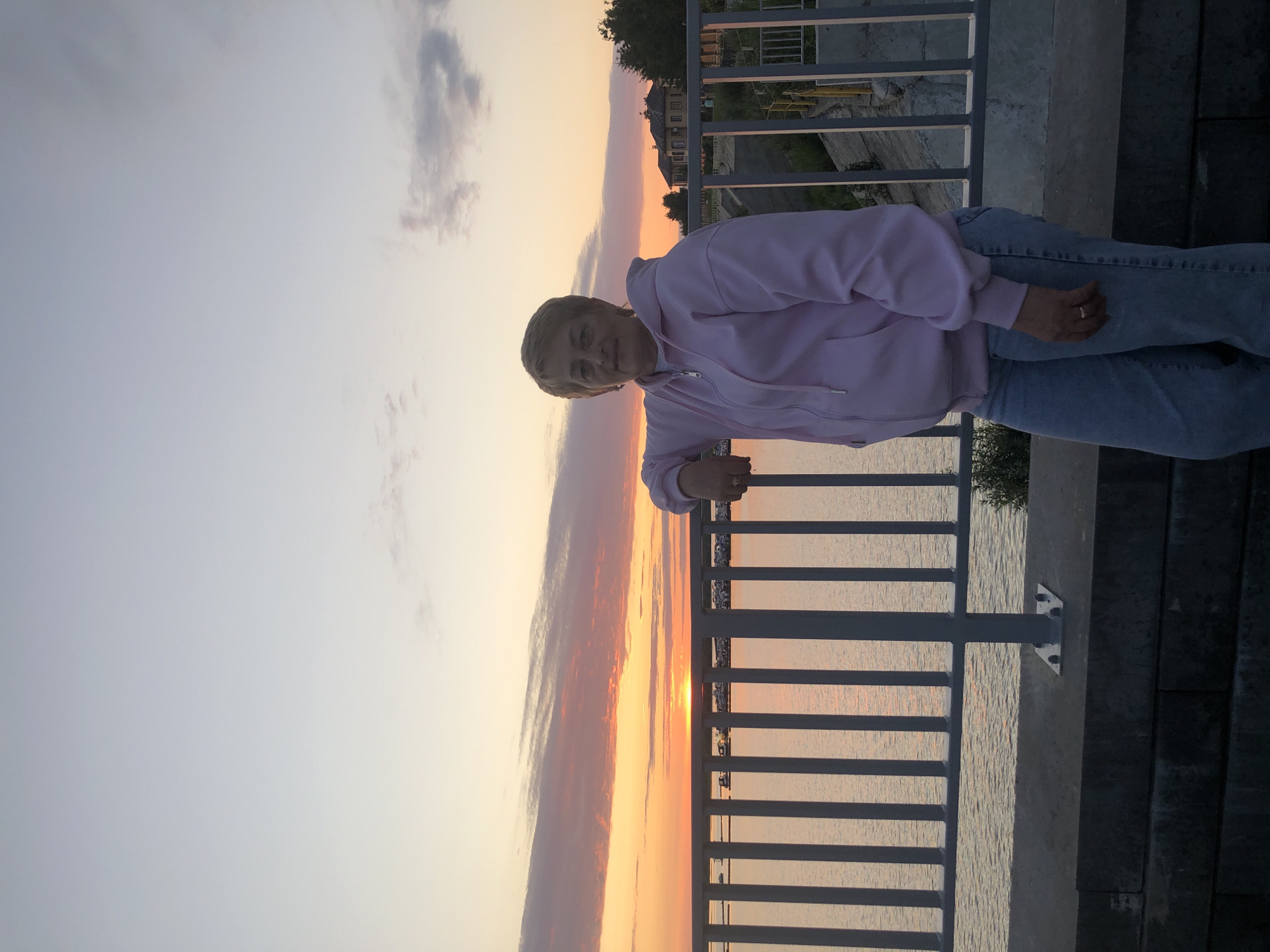 An elderly woman with short blonde hair stands on a bridge, leaning on the railing. She is wearing a light pink zip-up hoodie and light blue jeans. Behind her, the sky is painted with the warm colors of a sunset, with hues of orange and pink reflected on the calm water. The water appears to be a river or a large body of water, possibly the Kama River, given the location of Solikamsk, Russia. On the opposite bank, visible through the railing, are some houses and trees. The overall scene suggests a peaceful evening by the water.