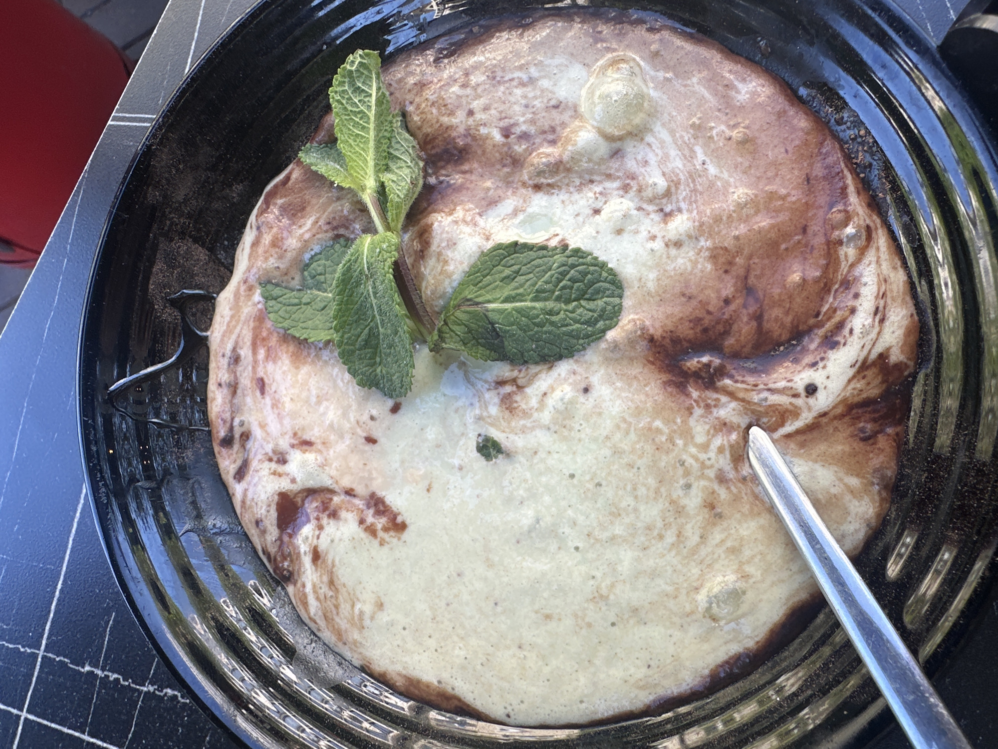 A top-down shot showcases a large, round pancake in a black pan, garnished with a sprig of mint. The pancake has a light brown and cream-colored marbled appearance. A silver fork rests on the right side of the pan, suggesting someone is about to eat the pancake. The pan sits on a black surface with white grid lines, possibly a tabletop or tray. A red object is partially visible in the upper left corner. The location is not identifiable as Sofia, Bulgaria from the image alone.