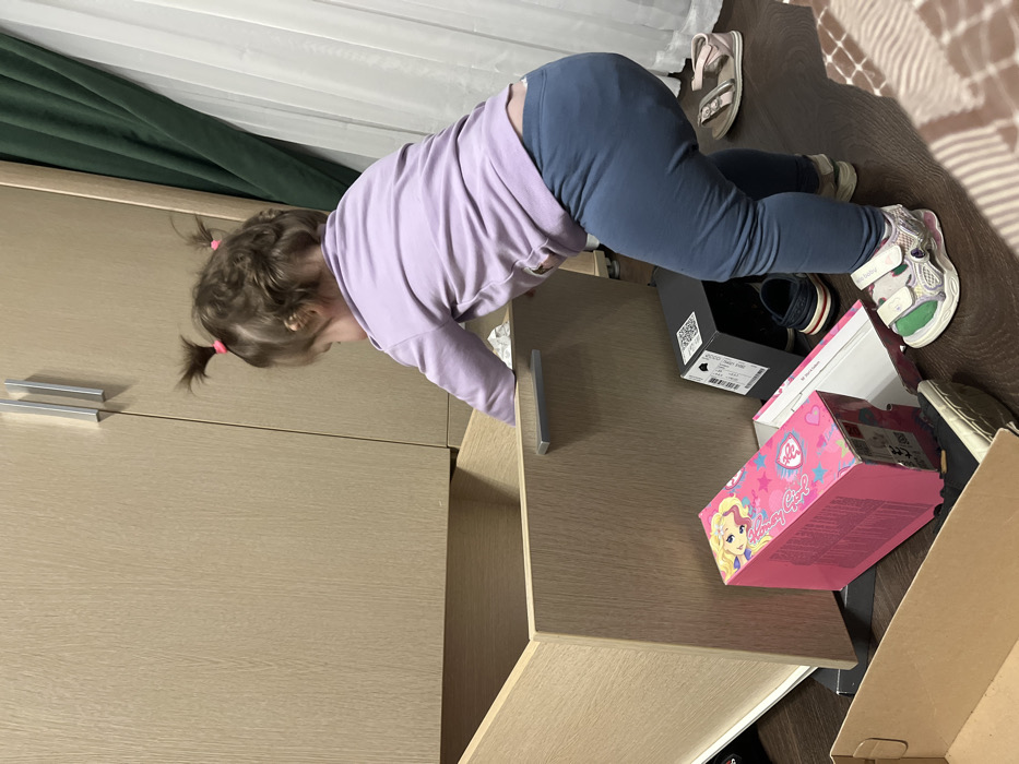A child with pigtails, wearing a lavender shirt and blue pants, leans over an open dresser drawer. The drawer is cluttered with shoe boxes, including a pink "Fancy Girl" box, and shoes. A pair of sandals and another pair of children's shoes are visible on the floor near the drawer. Green curtains hang in the background.