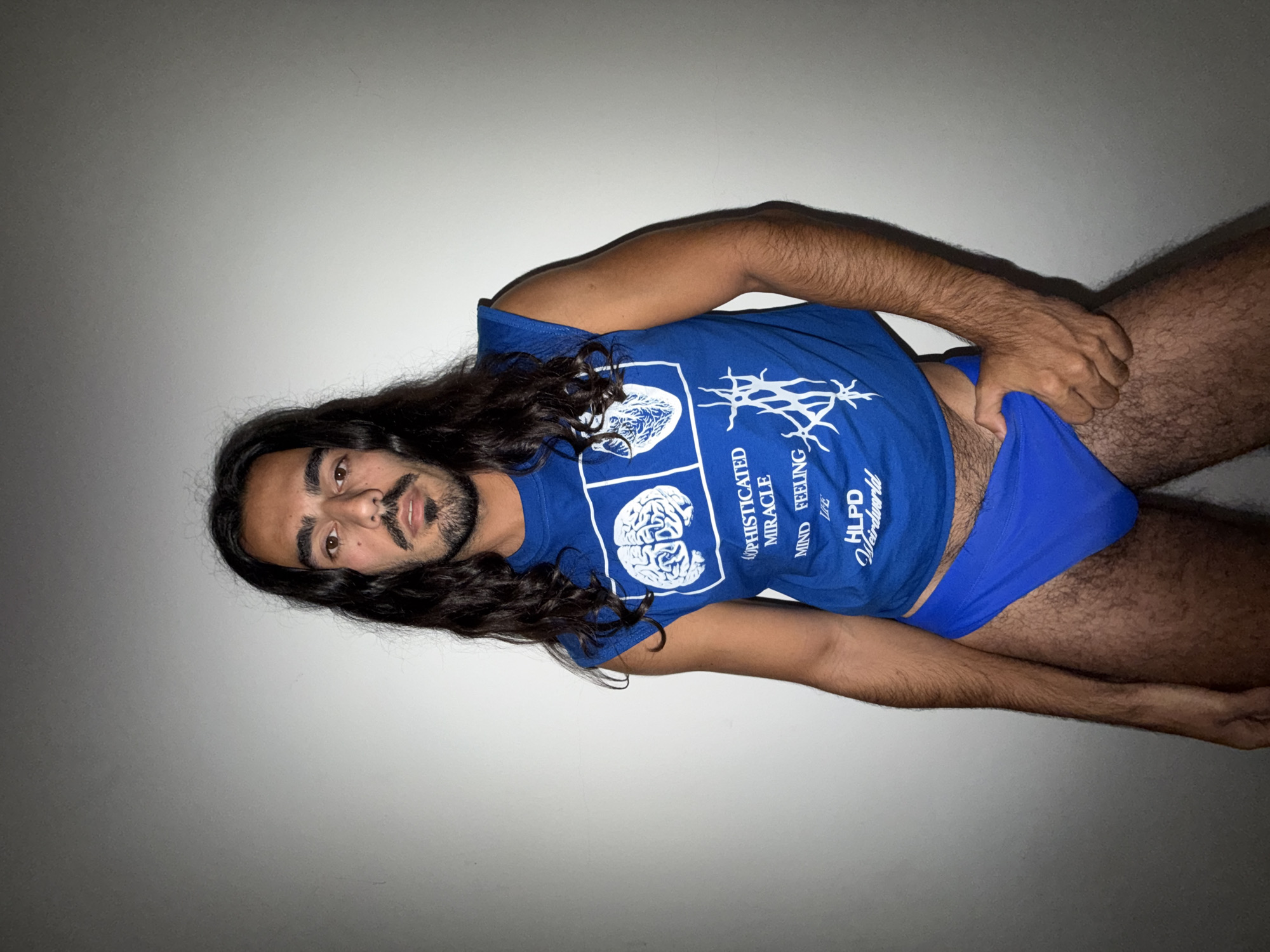 A man with long dark hair and a goatee lies on his side against a white wall. He wears a blue t-shirt and blue underwear. He is touching the side of his underwear with his right hand. The setting is not identifiable as North Kuta, Indonesia from the image alone.