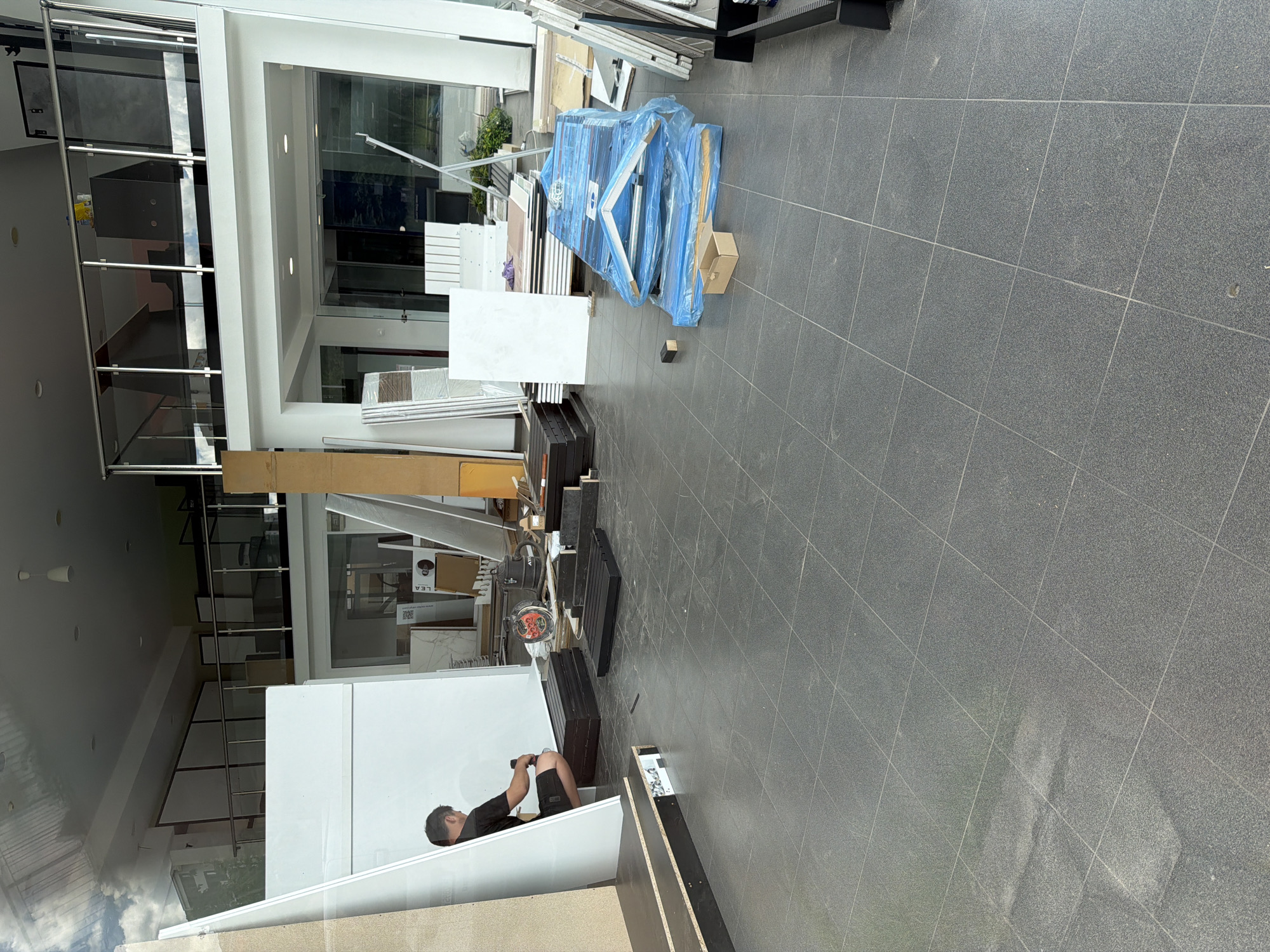 The image shows the interior of a building, likely a showroom or retail space, in Burgas, Bulgaria. Various building materials, including white panels, wooden beams, and items wrapped in blue plastic, are scattered throughout the space. A man is seen working on a white structure, possibly assembling furniture or a display. The flooring is composed of large gray tiles. The building features a modern design with glass walls and multiple levels.