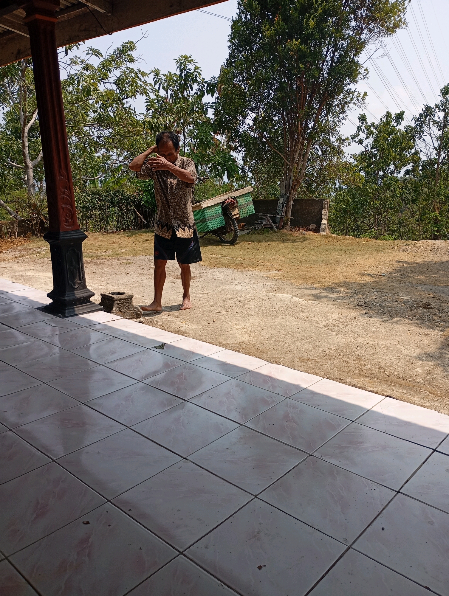 An elderly man wearing a patterned short-sleeved shirt and dark shorts stands barefoot on a dirt path in front of a tiled porch. He is facing away from the camera, with his arms raised, and appears to be wiping his face or adjusting his hair. Behind him, a motorcycle with a green basket is parked near a small, rough-hewn concrete structure and a tree. The scene is outdoors, with dry grass and various trees surrounding the area. The sky is visible above, and power lines are strung across the top of the frame. The overall impression is one of a rural or semi-rural setting in Ketepung, Indonesia.