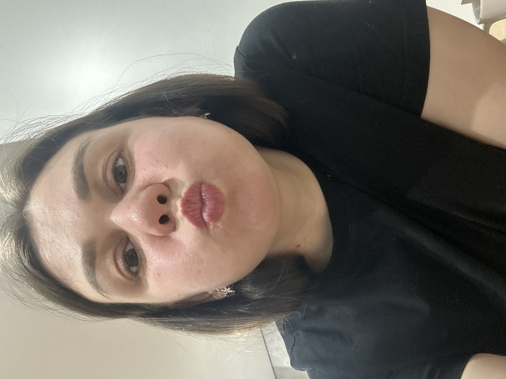 A woman with brown hair and brown eyes is looking at the camera with pursed lips. She is wearing a black shirt and earrings. The background is a plain white wall. It is not possible to determine the location as Chaikovsky, Russia with the provided information.