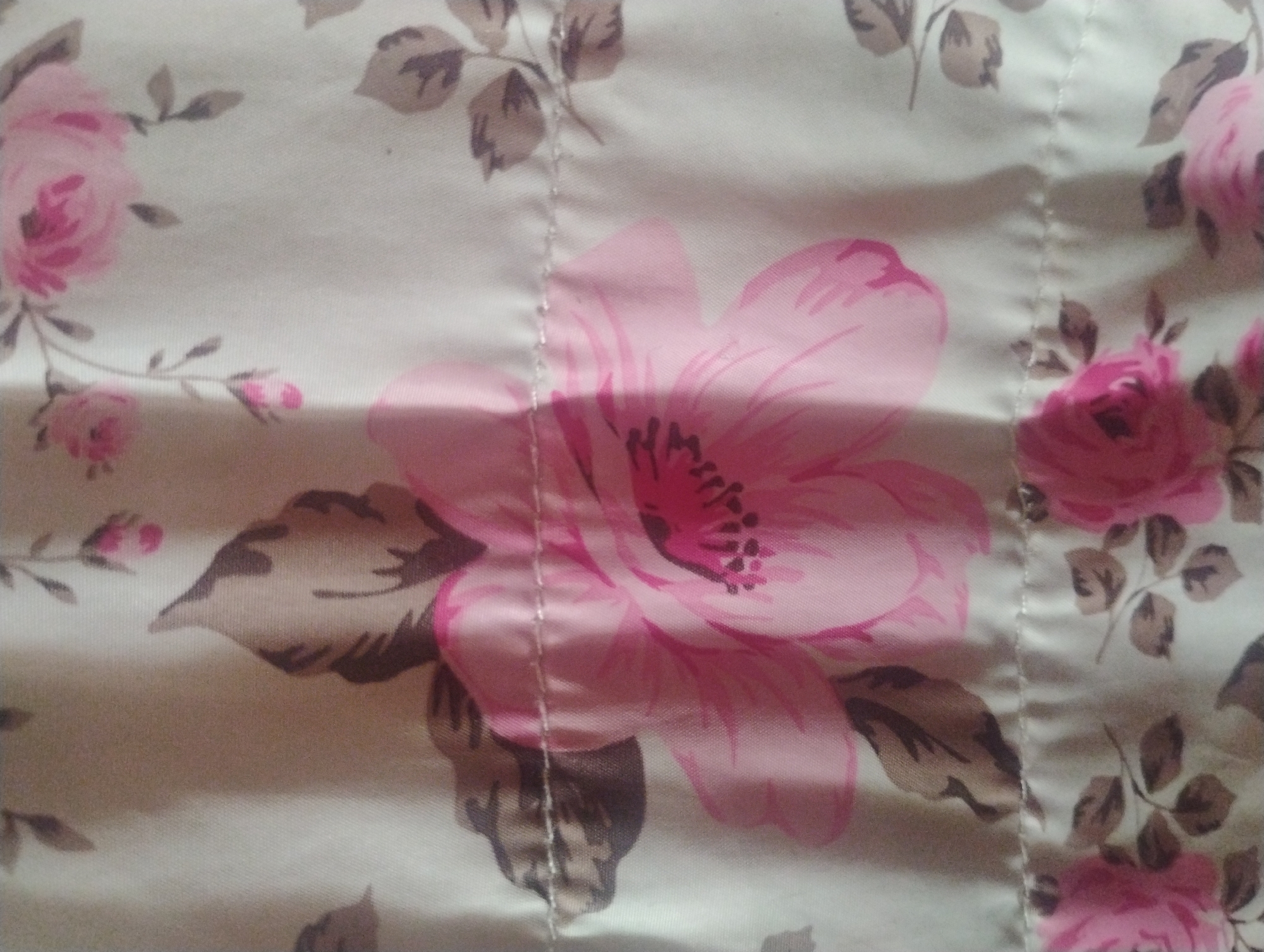 This is a close-up, high-angle photograph of a fabric with a floral pattern of pink roses and brown leaves. The fabric appears to be a light cream or off-white color. There is a visible seam running horizontally across the image, approximately in the center, with visible stitching. The lighting in the image creates some shadows and highlights on the fabric, giving it a slightly textured appearance. The image does not depict any part of the city of Rumyantsevo, Bulgaria, nor does it contain any people, actions, or interactions.