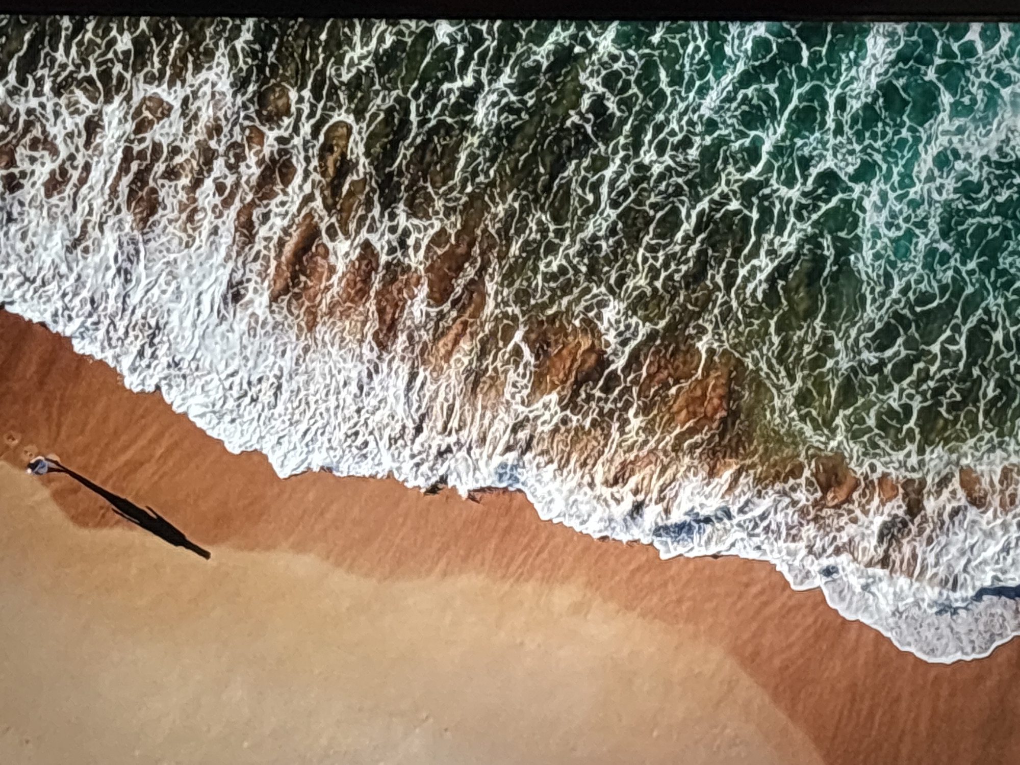 This is an aerial, high-angle, long shot of a person casting a long shadow on a sandy beach next to the ocean. The person is walking towards the left side of the frame. The ocean water is a deep blue-green with white foam from the waves crashing. The beach is a light brown sand color. The image is sharp and clear, with good lighting and detail. The location is not in Valladolid, Spain, as Valladolid is an inland city with no coastline.