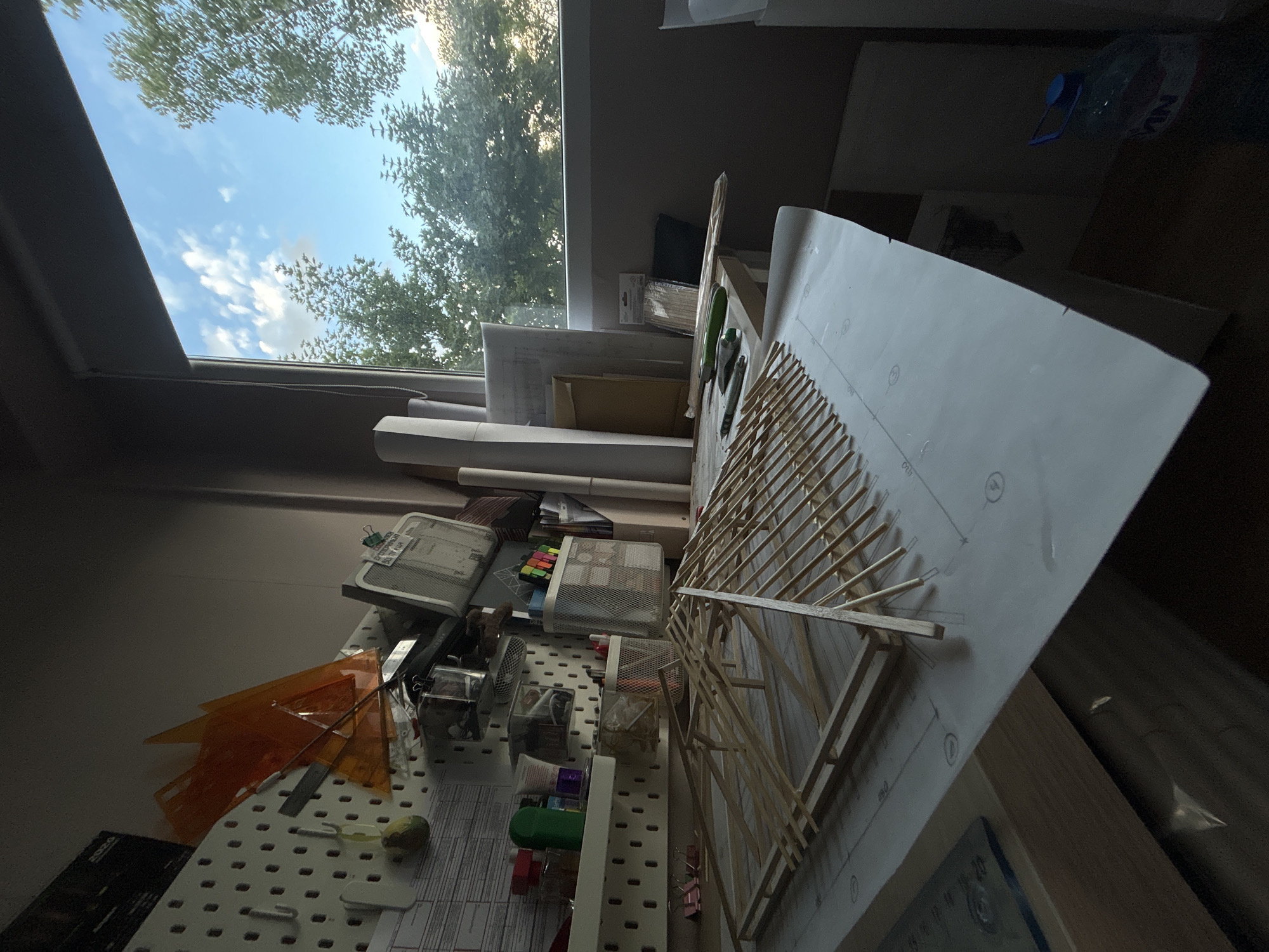 The image captures a vibrant, sunlit indoor workspace, likely a studio or design office in Sofia, Bulgaria, viewed from a slightly elevated, tilted perspective. The focal point is a desk or workbench covered with various creative and technical supplies.
Prominently featured is a skeletal model structure, constructed from numerous thin wooden sticks, possibly an architectural prototype or engineering design, resting on a large sheet of paper. This paper displays technical drawings, circular markings, and numbers such as "80" and "100", with some visible letter "D" markings.
To the left of the model, a white pegboard organizes an assortment of tools and materials. These include bright orange transparent set squares or triangles, rulers, and several mesh metal organizers filled with pens, pencils, and other small craft items. Below the pegboard, a calendar or planner displaying "2024" is visible. Further left, several rolled-up paper sheets, suggestive of blueprints or plans, are stacked. In the upper right corner of the desk area, a plastic water bottle with a blue cap shows partially visible letters "M" and "N" from its label.
A large window, possibly a skylight, dominates the upper left portion of the image, revealing a bright blue sky with scattered white clouds. Lush green tree canopies are visible outside the window, with sunlight filtering through the leaves.
The scene implies an ongoing creative or technical activity, such as architectural design or model-making. The bright daylight streaming through the window suggests the time of day is sunny morning or early afternoon. The overall impression is one of an organized, productive environment.