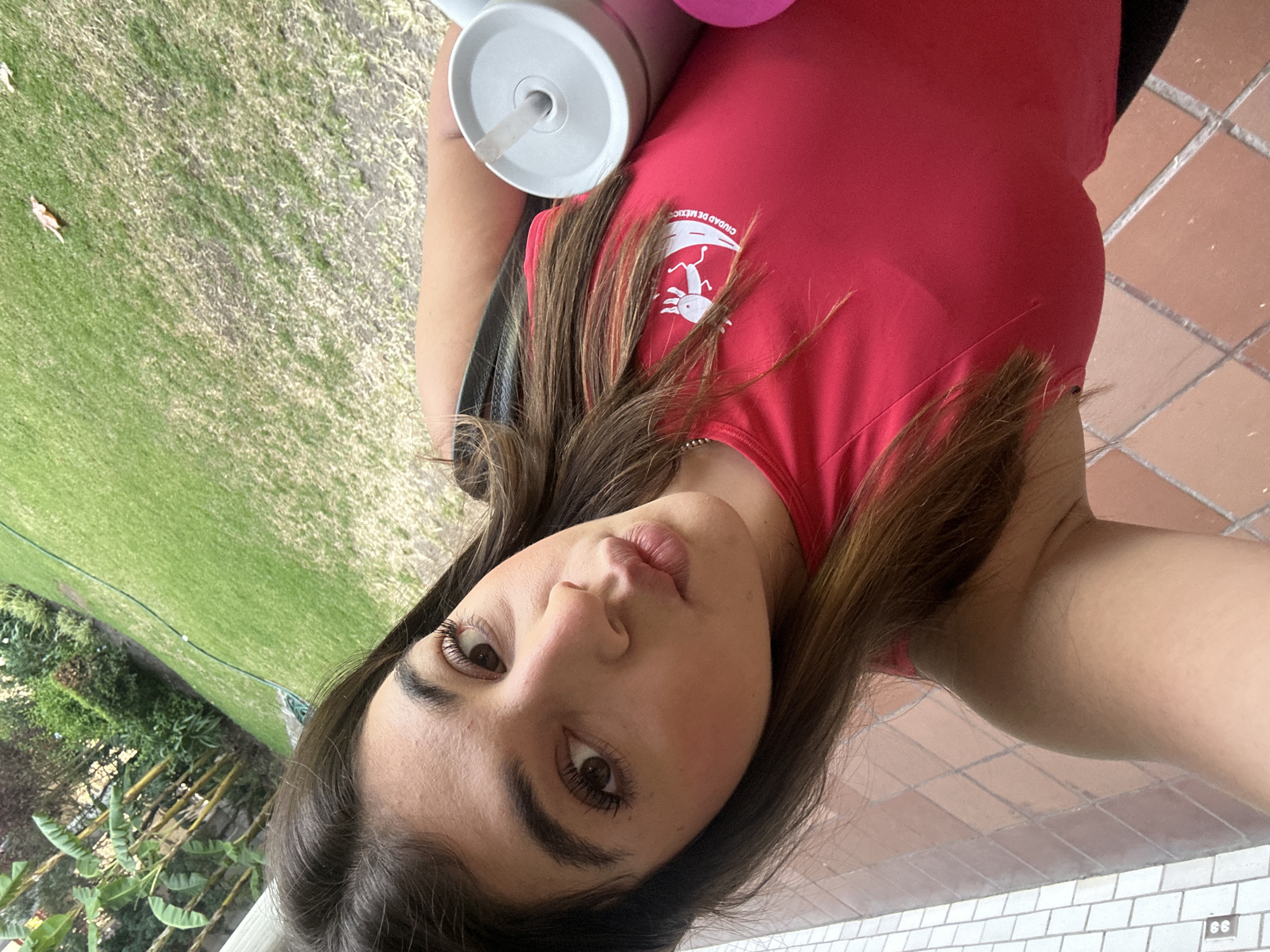 A woman takes a selfie in what appears to be an outdoor setting, perhaps a patio or backyard. She has brown hair, brown eyes, and is wearing a red shirt. The shirt has a logo on the upper left that includes a white, stylized salamander and the words "CIUDAD DE MEXICO". A drinking cup with a straw is visible next to her. The background includes a grassy area with some greenery, and a tiled patio surface. There is no indication of the specific location of San Bernardino Tlaxcalancingo, Mexico.
