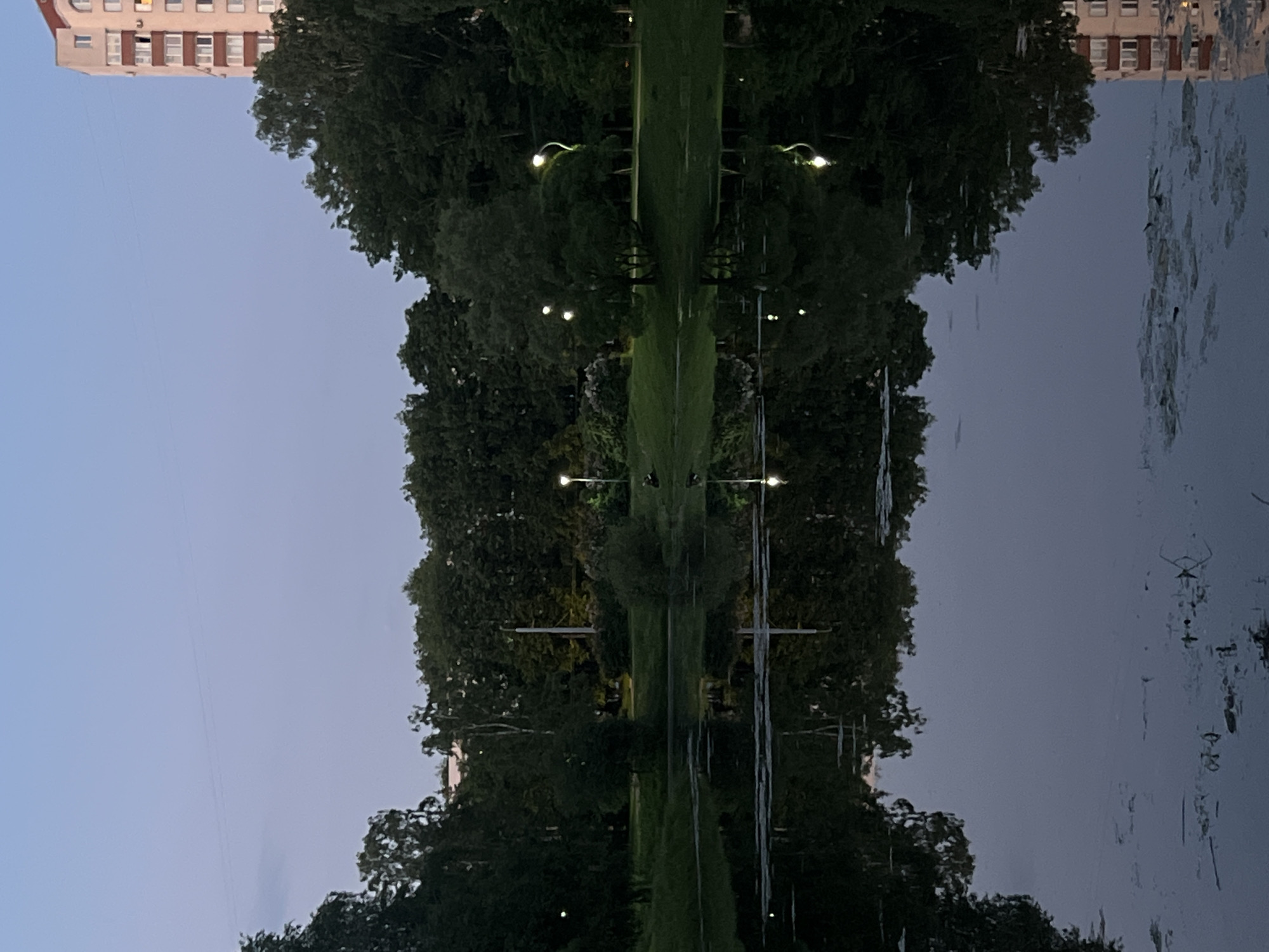 This image depicts a twilight or early night scene in a park-like environment within the city of Unknown, Russia. A calm body of water, likely a pond or lake, occupies the lower half of the frame, extensively reflecting the sky, surrounding trees, artificial lights, and a distant building.
The upper portion of the image shows a pale blue-grey sky, indicating low ambient light. Faint horizontal lines, possibly power lines, are visible across the sky.
The midground features a park with dense green deciduous trees bordering the water's edge. Several streetlights, some illuminated with a warm white glow, are positioned within the park, casting light on the grassy areas and creating clear reflections in the water. On the left-center bank, a dark silhouette of at least one person is seated on the grass near the water, with another dark, indistinct shape nearby.
In the background, to the right, a multi-story residential building with light-colored facades and a reddish-brown roof/trim is prominent. One window on an upper floor emits a distinct orange-yellow light. A smaller building is partially visible behind trees in the left-center background.
The water's surface in the foreground contains patches of floating aquatic vegetation. The reflections are clear and largely undisturbed, with the bright streetlights appearing as elongated streaks of light. No specific actions beyond the presumed resting of the seated individual(s) are discernible.