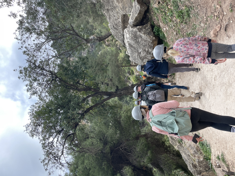 A group of people, equipped with light grey helmets, are walking along a dirt or gravel path through a rugged, natural landscape in El Chorro, Spain. The scene suggests an outdoor adventure activity like hiking or trekking, possibly along a challenging route, given the safety helmets.
In the foreground, with her back to the camera, a person wears a light pink long-sleeved top, black leggings with white stripes on the left calf, and a light green backpack. She holds a dark-colored smartphone in her left hand. Directly behind her, another person wears a dark grey backpack, khaki pants, and a dark top. Further back, more individuals are visible, also wearing helmets and varying outdoor attire, including a dark blue jacket and jeans, and another in a blue jacket with dark trousers. Towards the right side of the path, a person with blonde hair is seen from behind, wearing a floral-patterned jacket with pink, white, and black colors, and black and white checkered pants.
The path is flanked by large, grey rock formations on the right and dense green trees and foliage on the left. The terrain appears uneven and rocky off the path. The sky above is overcast with soft, diffuse light, indicating it is daytime. The natural setting features various types of trees, some with visible branches against the sky, while others are full of leaves, contributing to a lush environment within the rocky gorge characteristic of El Chorro. There is no visible text in the image.