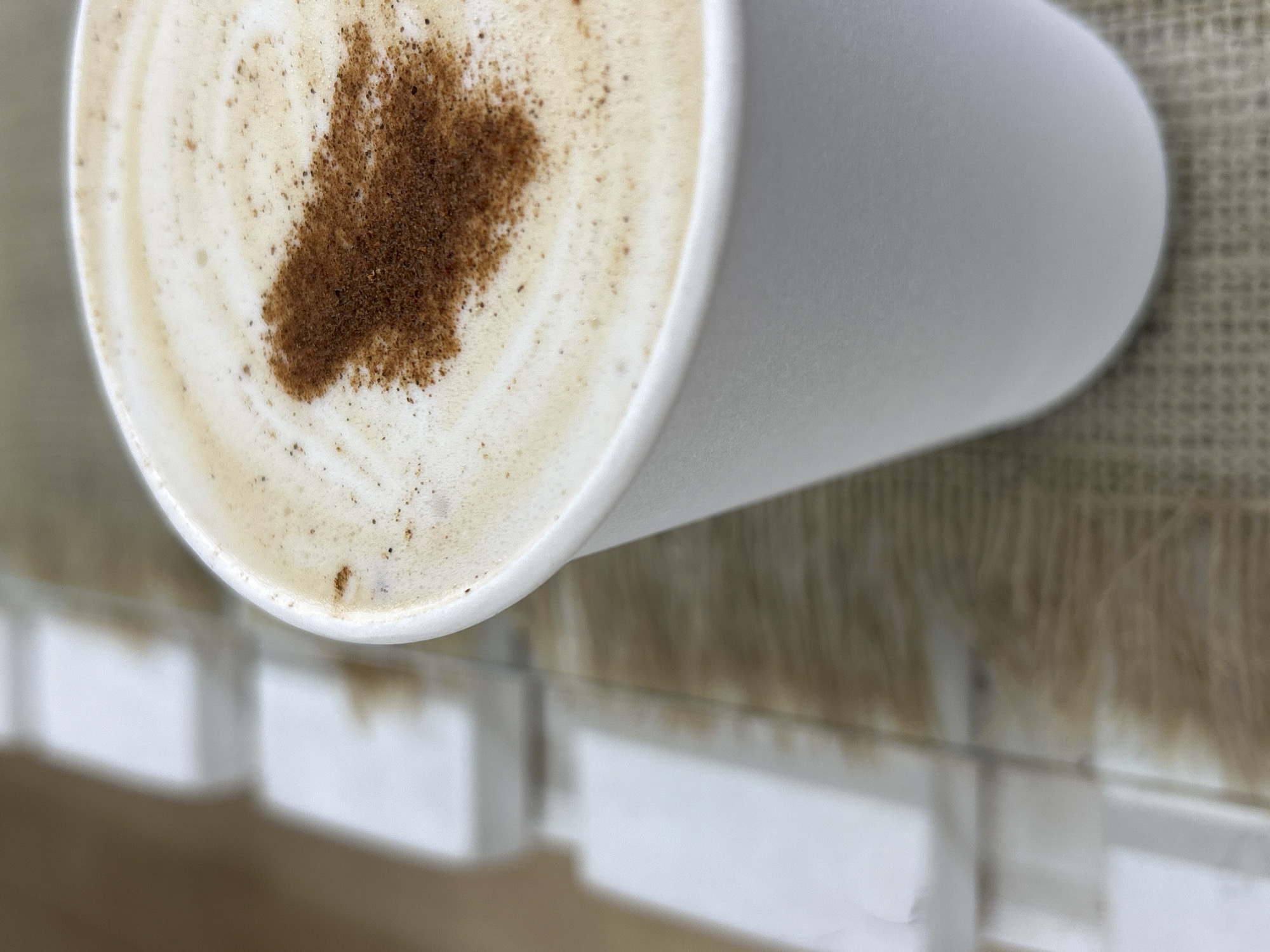 An aerial shot shows a white paper cup of coffee with milk foam, dusted with cinnamon, sitting on a white surface with a textured tan and white backdrop. The focus is sharpest on the coffee cup, with the background elements appearing blurred. No people are visible in the image. The location, Chaikovsky, Russia, is not readily discernible from the visual elements present.