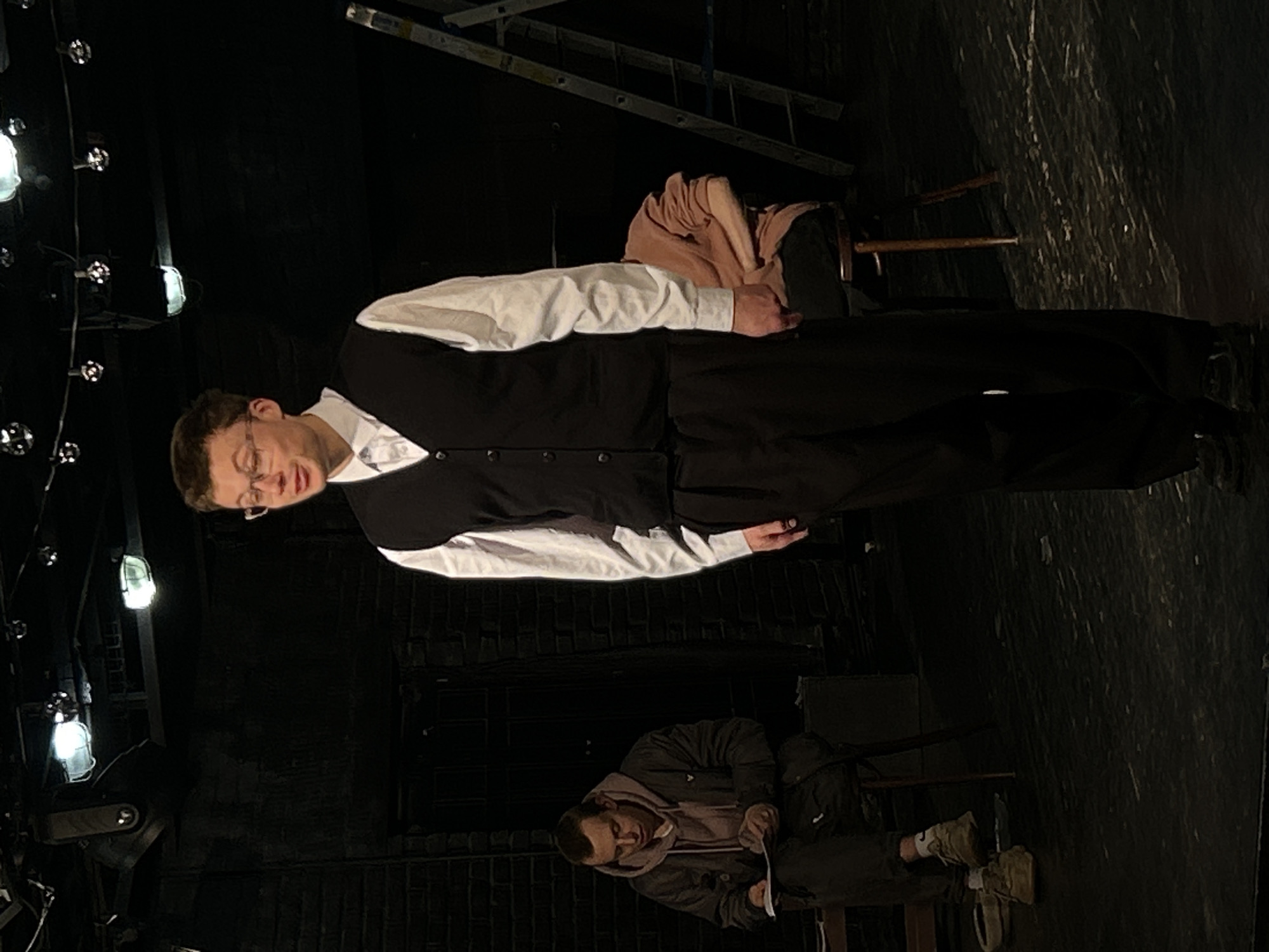 The image depicts two men in what appears to be a theatrical stage or rehearsal setting, characterized by dim lighting and stage equipment.
In the foreground, a man stands dressed in a white long-sleeved shirt, a black button-up vest, black trousers, and glasses. His head is tilted downwards and slightly to his left, with his eyes cast in the same direction, suggesting a focused or pensive expression. His left arm is bent, and his right arm hangs alongside his body. Behind him, to his right, a light brown fabric is draped over the back of a chair.
Further back in the scene, another man is seated on a chair. He wears a dark jacket over a lighter hoodie and is engrossed in examining a piece of paper or a script held in his hands.
The left side of the frame features a string of bare lightbulbs, some illuminated, running vertically, and what appear to be stage spotlights or fresnels mounted on stands or pipes. In the upper background, a long, light-colored ladder rests horizontally, possibly part of a set or stage rigging. The floor is dark and has a textured appearance. The overall scene suggests a rehearsal or preparation for a performance. No visible text is present.