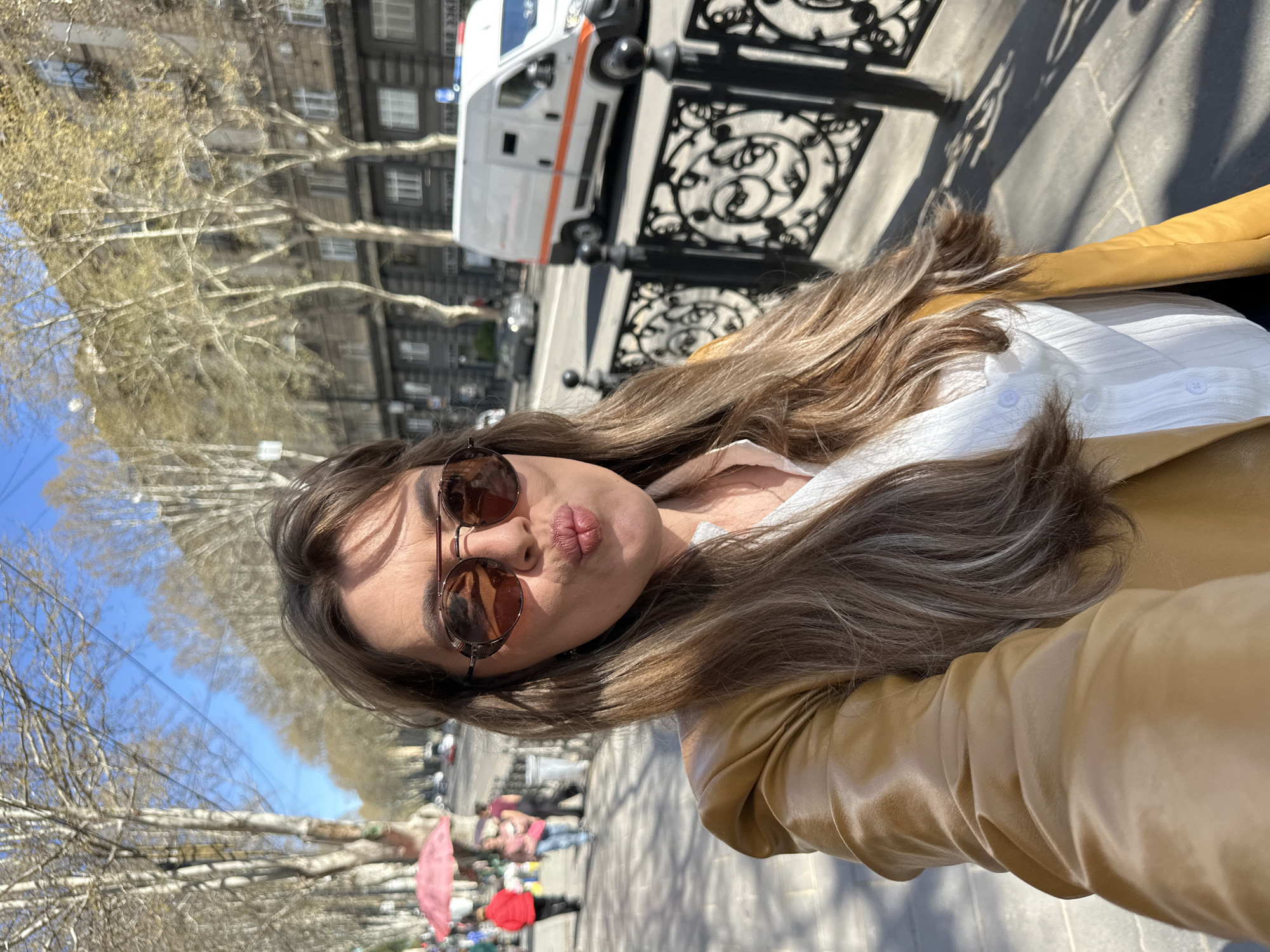 A woman in sunglasses and a tan jacket takes a selfie outdoors. She has long brown hair and wears a white shirt under the jacket. The background shows a street with a line of trees and an ornate black metal fence. Across the street, there is a building and a white van. In the distance, people are visible. Based on the architecture and context, the location is likely Tbilisi, Georgia.