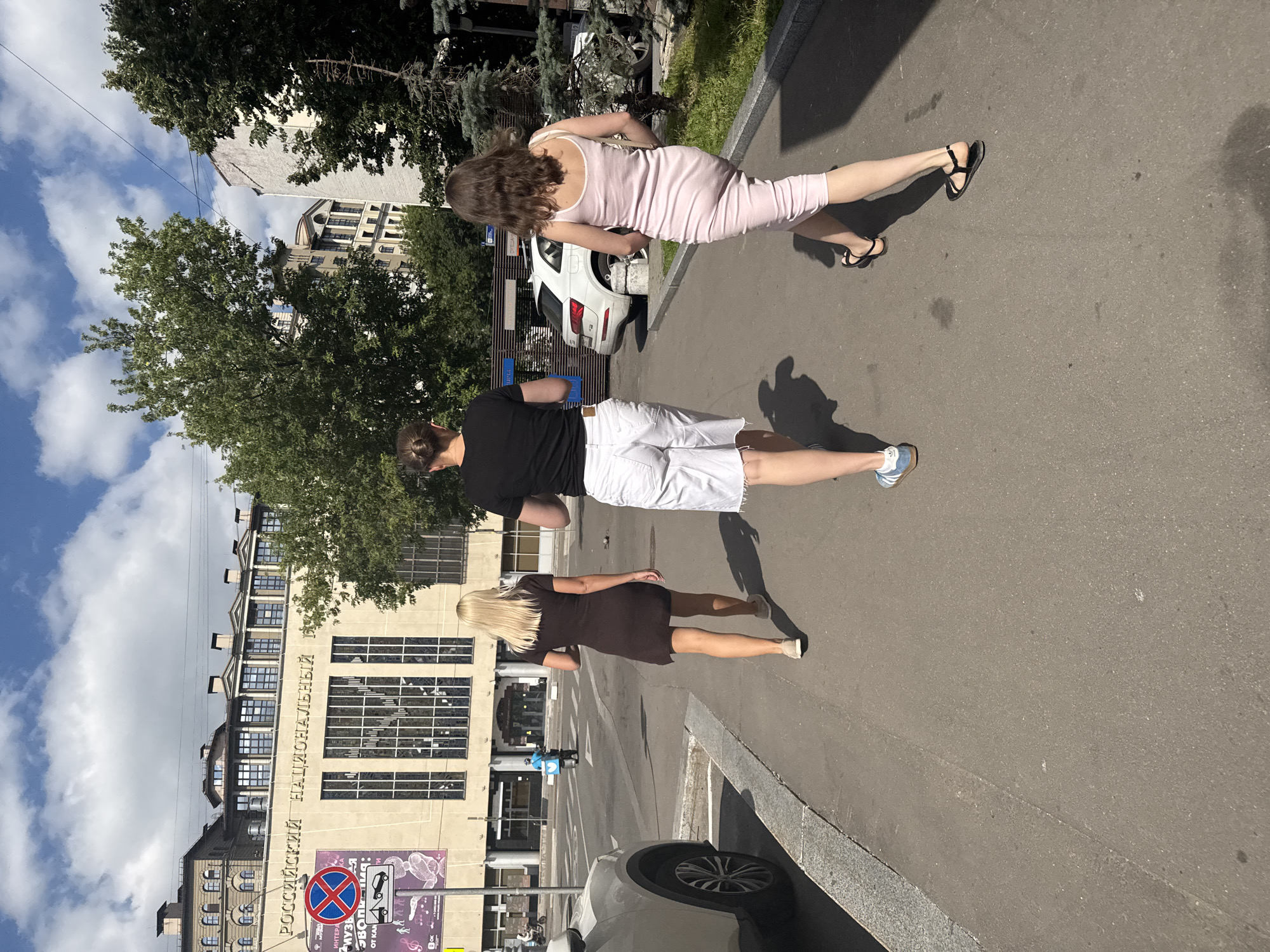 Three individuals are walking away from the camera on a sidewalk next to a street. The person in the middle, wearing a black t-shirt and white shorts, has their back to the camera and is walking ahead of the other two. To their left, a woman with blonde hair, wearing a dark brown dress, is walking slightly behind and to the side. To the right of the person in the middle, a woman with brown hair, wearing a pale pink dress, is walking parallel to the person in the middle.
The building behind them features a large sign that reads "РОССИЙСКИЙ НАЦИОНАЛЬНЫЙ" (Russian National). This is likely part of the building's name or a description of its purpose. In the background, there are other buildings, trees, a street with a white car, and a blue scooter with a delivery box. The sky is partly cloudy. This scene is located on a street in Moscow, Russia, given the Russian text on the building.