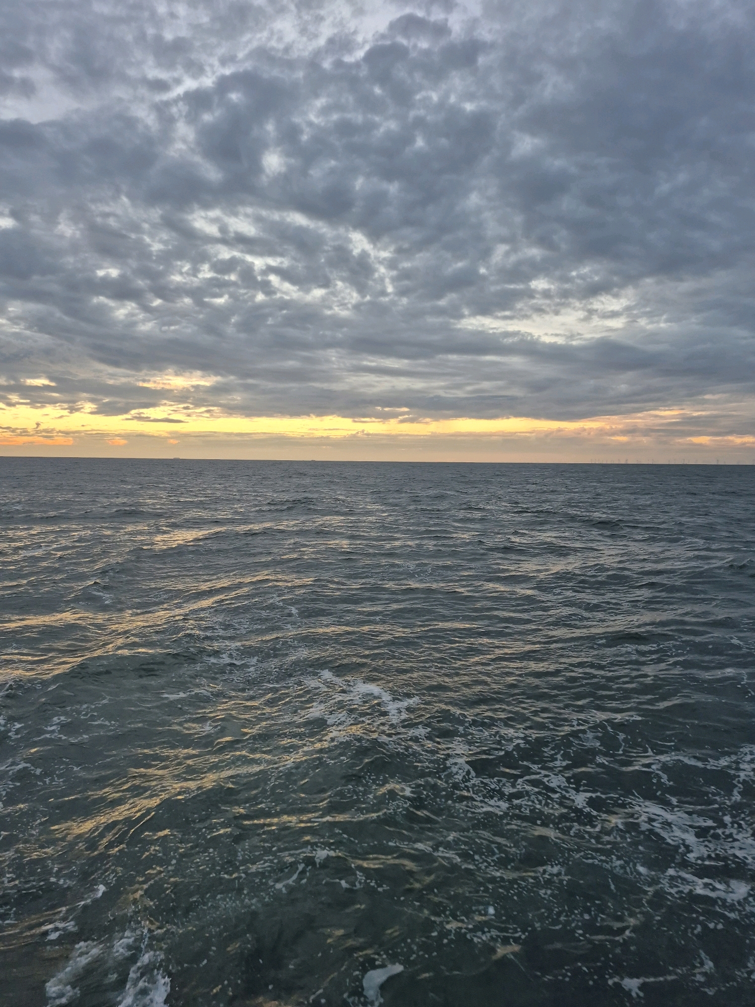 An image captures a cloudy sky over a dark, choppy sea. The horizon is faintly illuminated with a band of yellow and orange light, suggesting either sunrise or sunset. The sea is a deep gray-blue, with white foam cresting the waves. In the distance, silhouetted against the horizon, are several wind turbines, indicating an offshore wind farm. The image is devoid of human presence or any specific landmarks that would pinpoint a location within the city of Unknown, Germany. The environment depicted is one of open water under a moody sky.