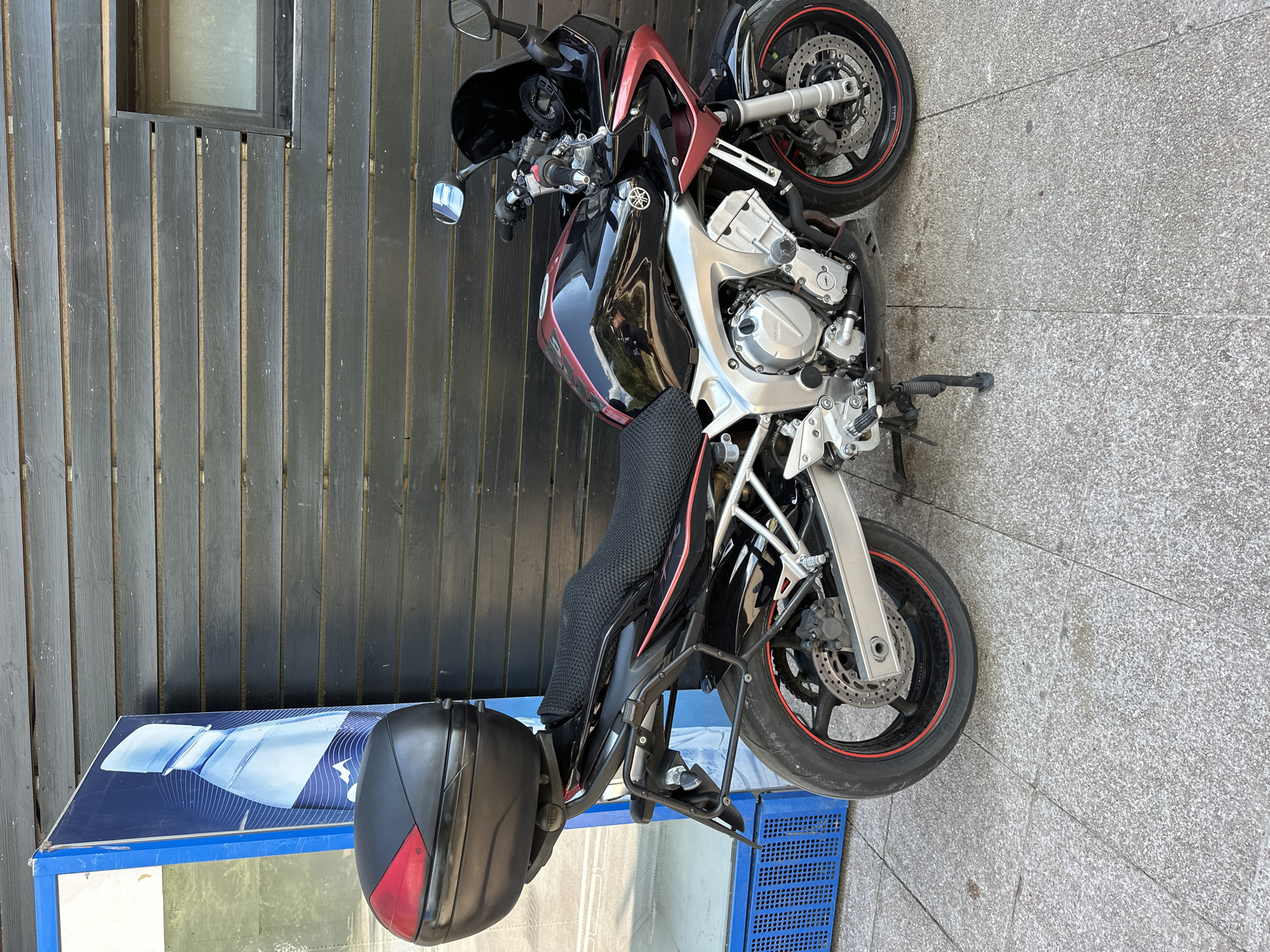 A black and red Yamaha motorcycle is parked on a grey tiled sidewalk. The motorcycle has a black top case with a red accent on the back and a textured black seat. It is positioned next to a blue structure with a large advertisement featuring a bottle of water. Behind the motorcycle is a building with dark wooden plank siding and a small, frosted window. The scene appears to be in an urban outdoor setting, possibly in Budva, Montenegro.