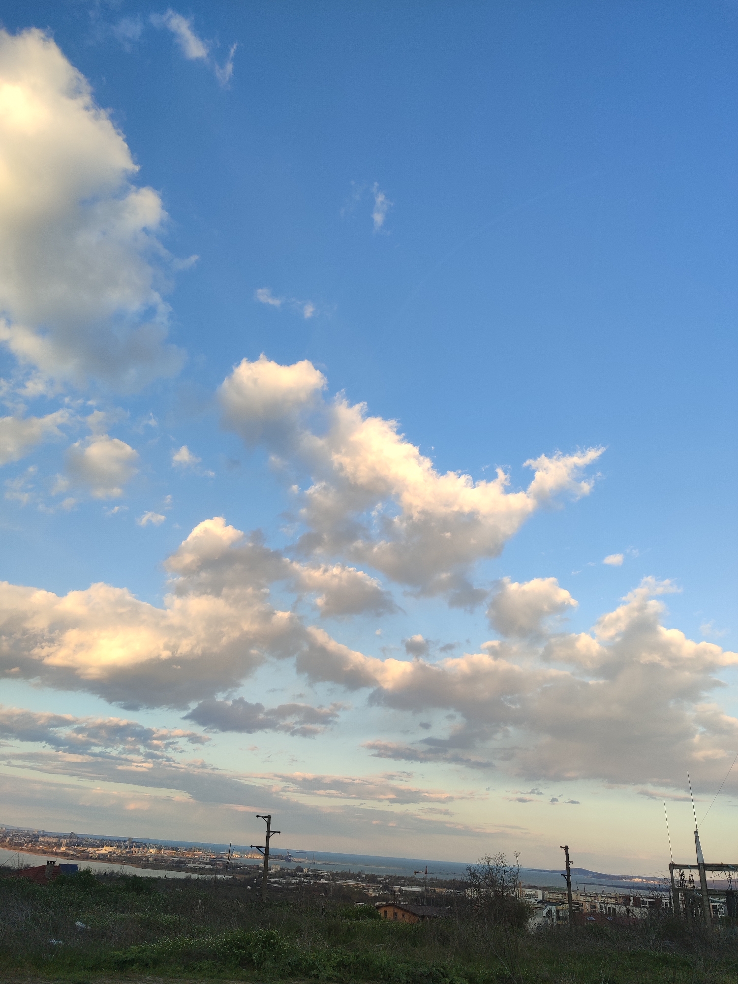 The image captures a wide-angle view of Burgas, Bulgaria, under a sky filled with scattered cumulus clouds illuminated by the sun. The sky transitions from a deep blue at the top to a lighter hue near the horizon.
The foreground shows a grassy slope with visible vegetation. In the midground, the cityscape of Burgas unfolds, featuring a mix of residential and industrial buildings, power lines, and glimpses of water bodies, possibly part of the Port of Burgas or the Black Sea. A faint, almost imperceptible circular arc (perhaps a contrail) is present high in the sky. No people are visible in the image.