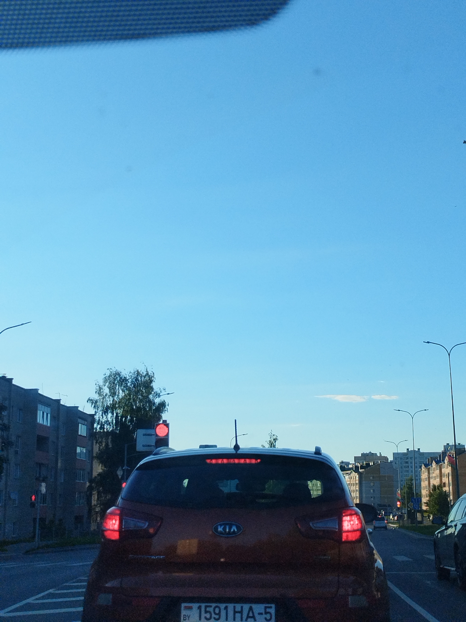 An eye-level shot captures a traffic scene on a clear, blue-sky day. In the foreground, the rear of a red Kia SUV is visible, its brake lights illuminated. The vehicle has a license plate with the text "BY 1591 HA-5". To the left, a multi-story apartment building stands, with windows and balconies. A traffic light with a red signal is positioned to the left of the SUV. Further down the road, other vehicles are seen in traffic. Streetlights and trees line the urban landscape. The image appears to be taken from inside another vehicle, looking forward.