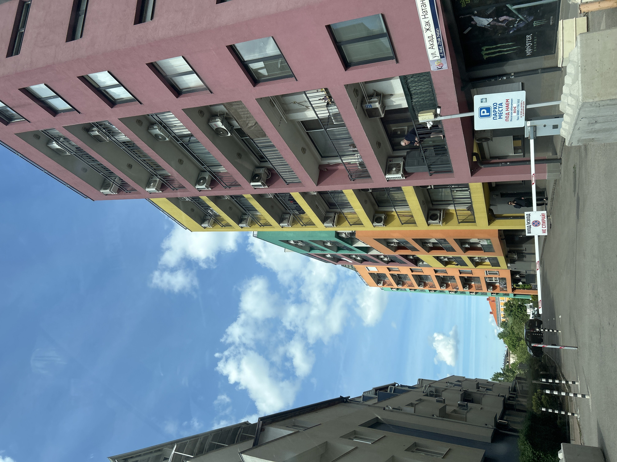 The image shows a view of buildings with colorful facades in Sofia, Bulgaria. The buildings are painted in shades of pink, yellow, green, and orange. Windows and air conditioning units are visible on the building exteriors. A street sign indicates "ul. Akad. Zhak Natan" (Acad. Zhak Natan street). A parking sign indicates "МЕСТА ПОД НАЕМ" (Spaces for Rent) with a monthly subscription of 80€. There is also a street and a gate with a stop sign that says "НЕ СПИРАЙ!" (Don't Stop). The sky is blue with clouds.
