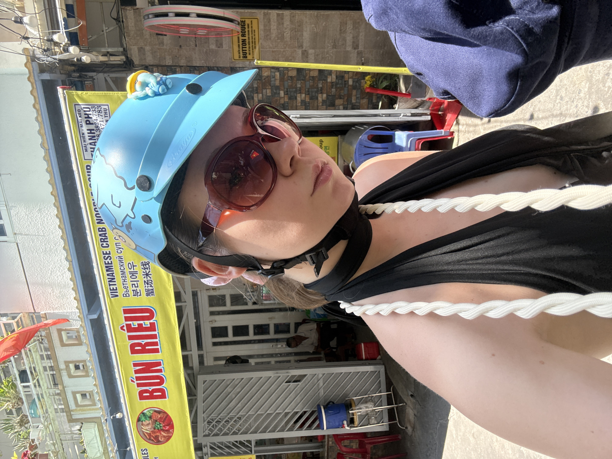 A medium shot captures a person with fair skin, wearing a blue helmet with cartoon-like designs and a yellow figurine on top. The person is also wearing sunglasses with a red frame and a black tank top. A white rope is draped around their neck.
Behind the person, a yellow banner displays the words "BÚN RIÊU" in large red letters. The banner also features text in Vietnamese, Russian, and Korean. A sign with Vietnamese text and the words "BUTTON ROUGE" is also visible.
The location appears to be a street in Nha Trang Ward, Vietnam. A red flag hangs in the background, and a building with white walls and windows can be seen behind the banner. A person is sitting inside the building. Various objects, including plastic chairs and water jugs, are scattered around.