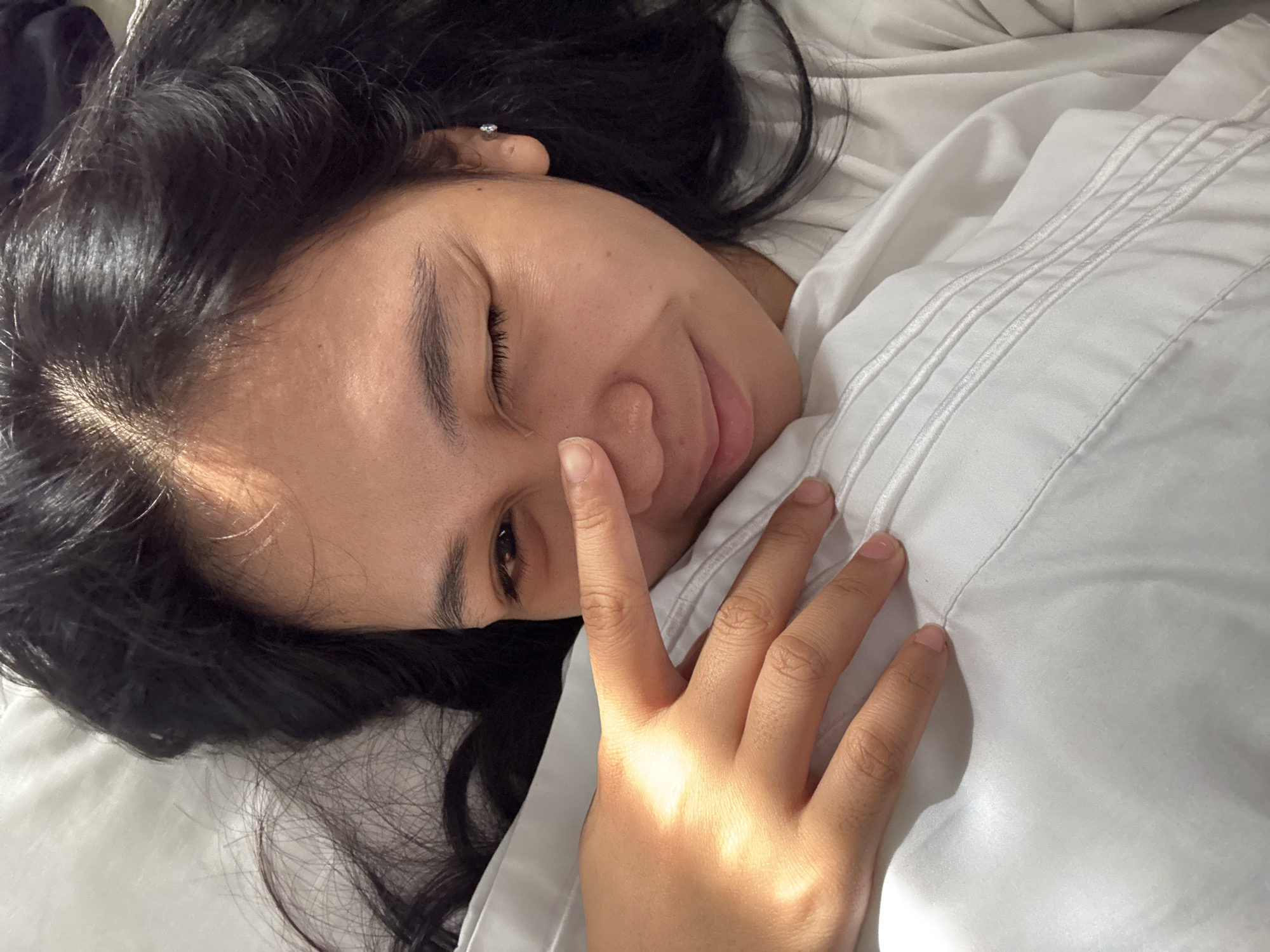 A high-angle, close-up image depicts an adult female, appearing to be of East/Southeast Asian descent, lying on a white surface, consistent with a bed or pillow, in an indoor setting located in Davao City, Philippines. The individual has long, dark hair spread around her head. Her right eye (viewer's right) is winked shut, and her lips form a slight, closed-mouth smile. Her left eye (viewer's left) is partially obscured by her raised left hand, with the index finger touching the bridge of her nose. A small, clear stud earring is visible in her left earlobe. Strong natural light illuminates the scene from the viewer's right, creating distinct highlights on her skin and the white fabric, which displays subtle texture and a pattern of parallel stitched lines.