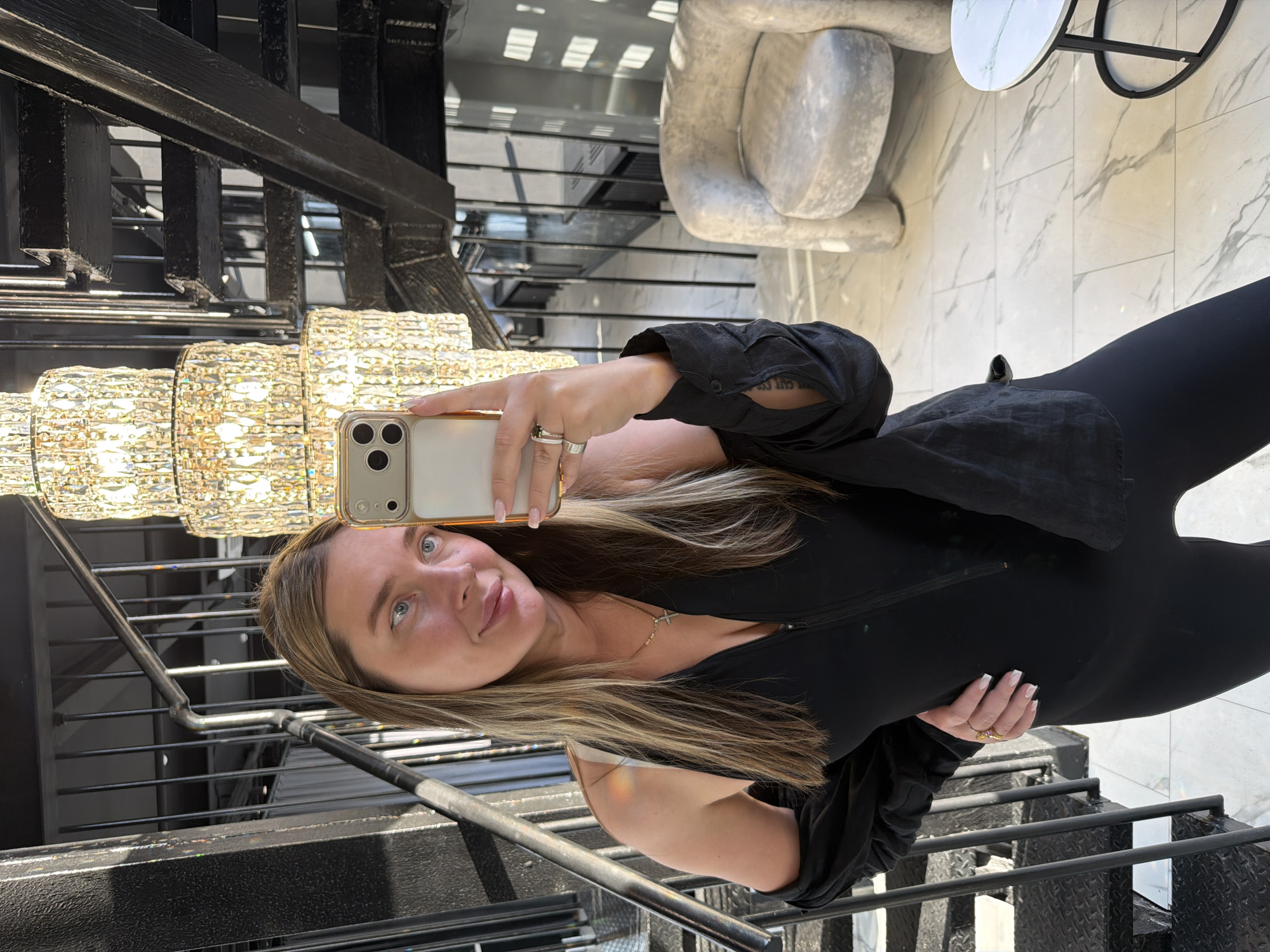 A woman is taking a selfie indoors, likely in a commercial building, standing by a metal staircase and a large crystal chandelier. The location, suggested by landmarks, is in Moscow, Russia.
The woman is wearing a black jumpsuit and a black, long-sleeved shirt, and holding a smartphone. She has light-brown hair with blonde highlights and is wearing rings on her fingers.
Behind her, the interior design features a modern aesthetic with marble floors and a white, abstract sculpture. Sunlight is entering through windows, creating highlights on the chandelier and staircase.